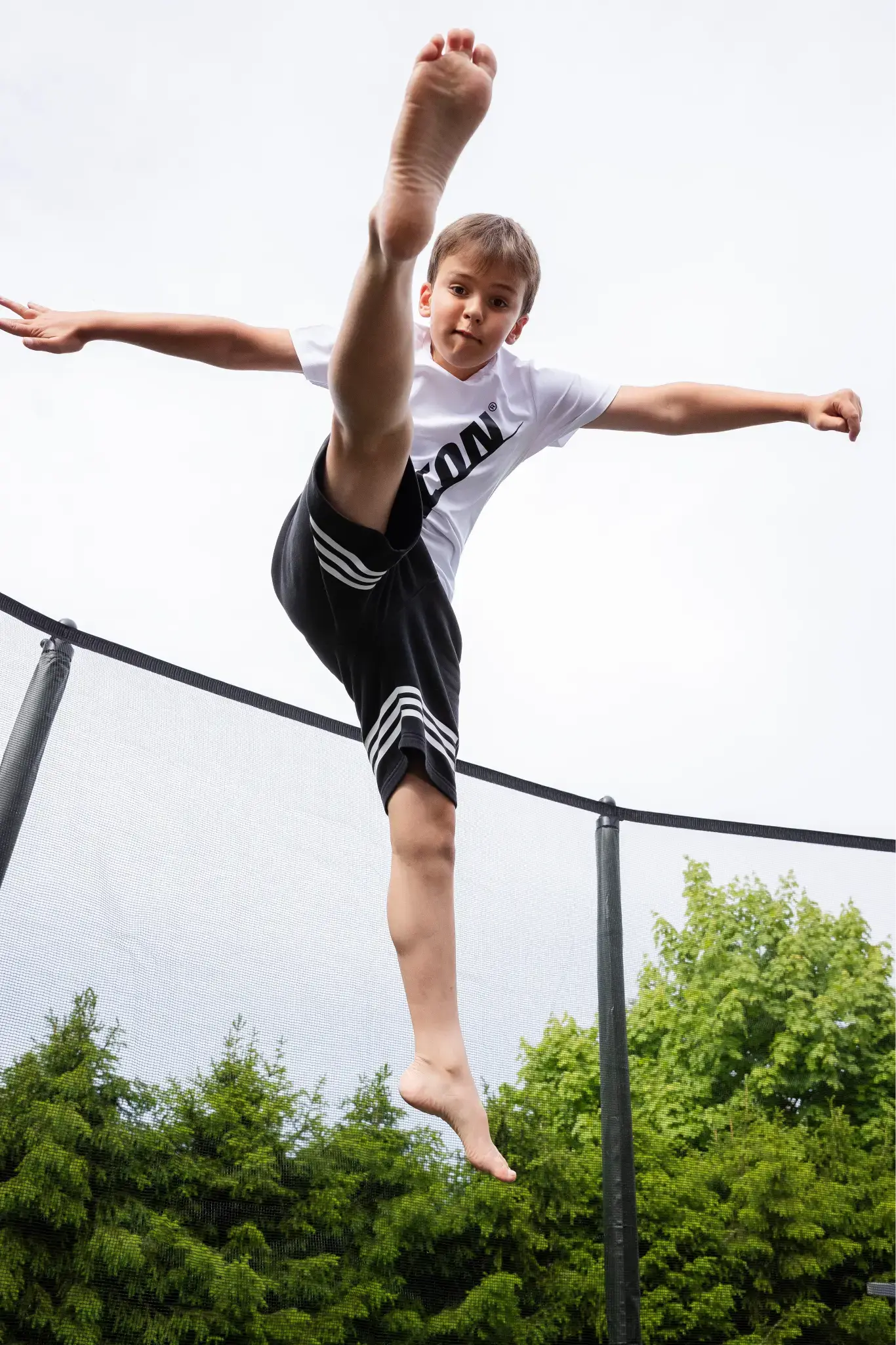 A boy performing a high kick on a trampoline, with one leg extended above his head. He is wearing a white Acon t-shirt. The background shows green trees and a cloudy sky."