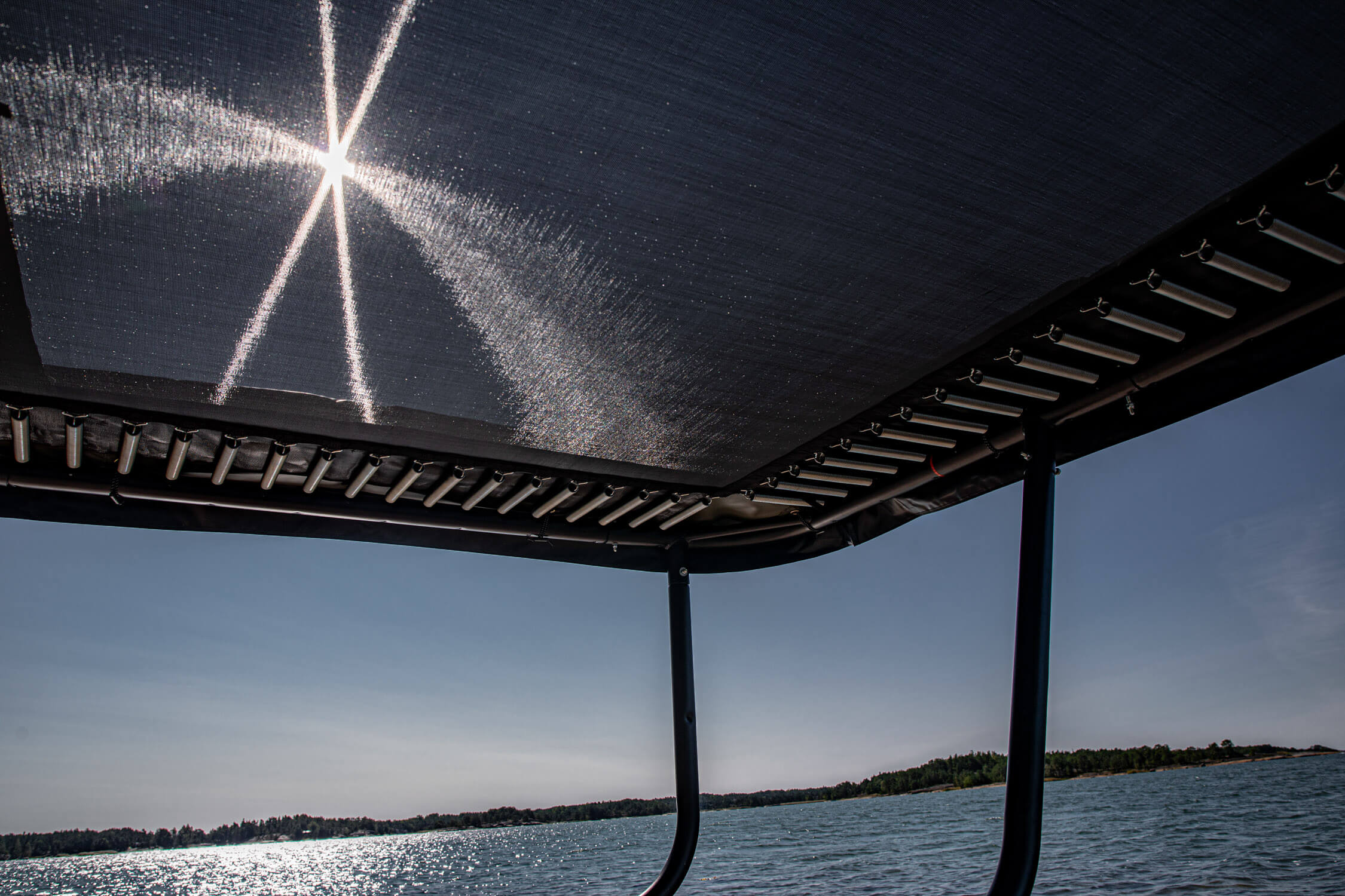 Acon trampoline from worm's eye perspective. The sun rays are hitting the trampoline mat. There's a lake in the background.