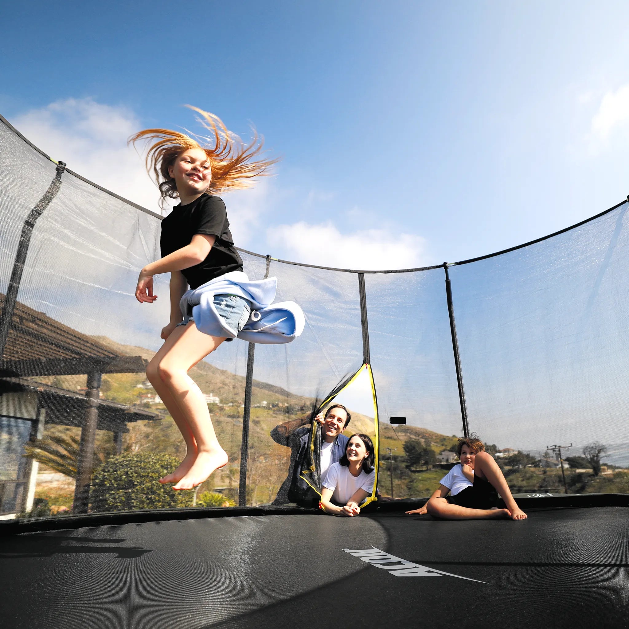 A happy girl is jumping on an Acon trampoline while her mother, father, and little brother watch.