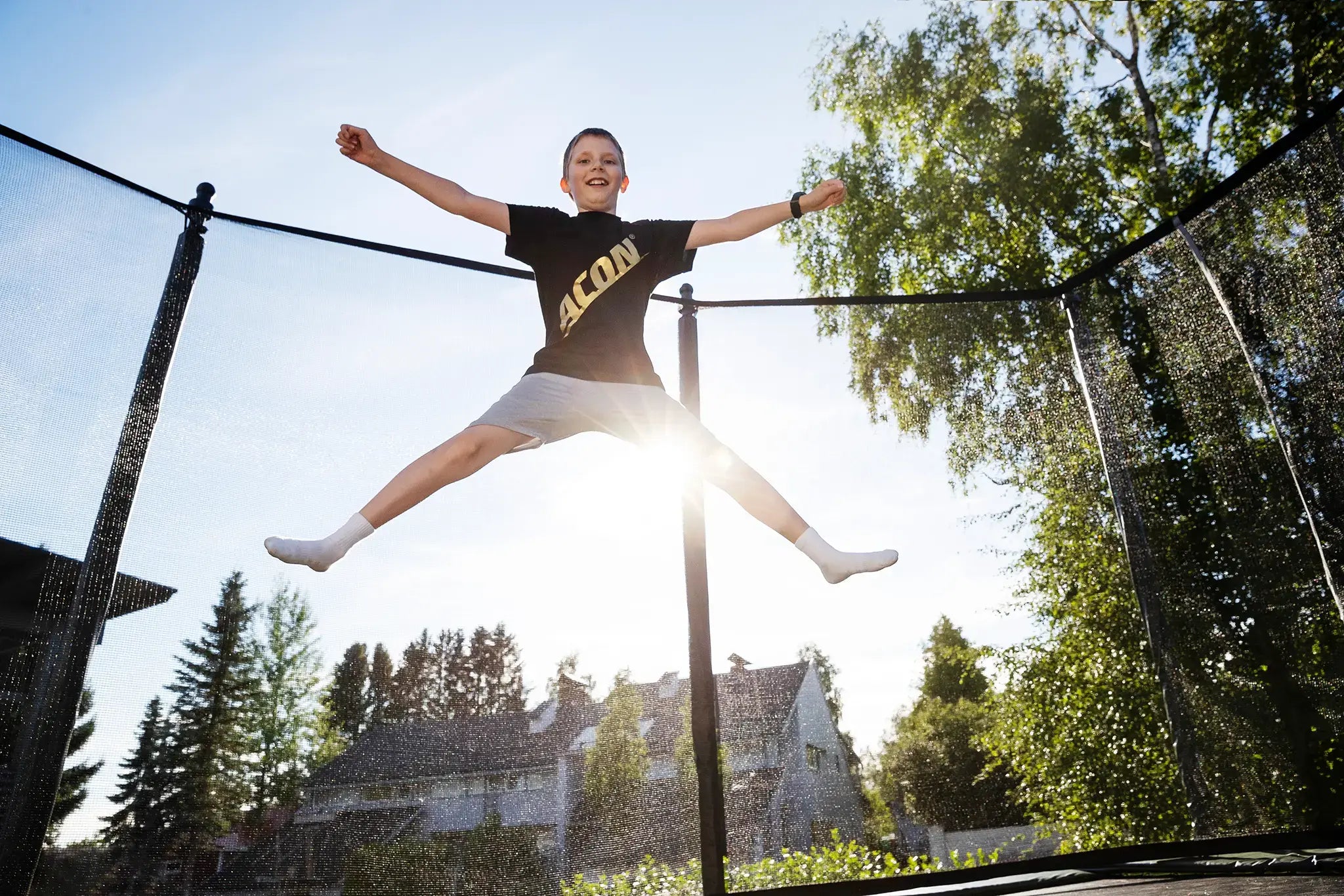 Boy wearing an Acon t-shirt while doing x-jump on an Acon trampoline, with sun shining in the backyard.
