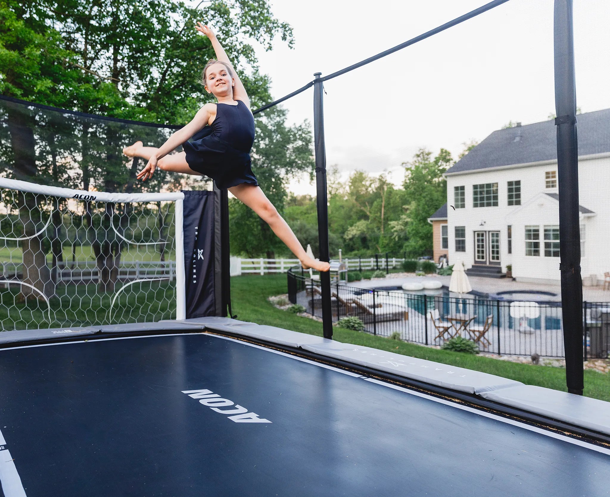 Girl jumps high on ACON trampoline in backyard, smiling mid-air near a house and pool.