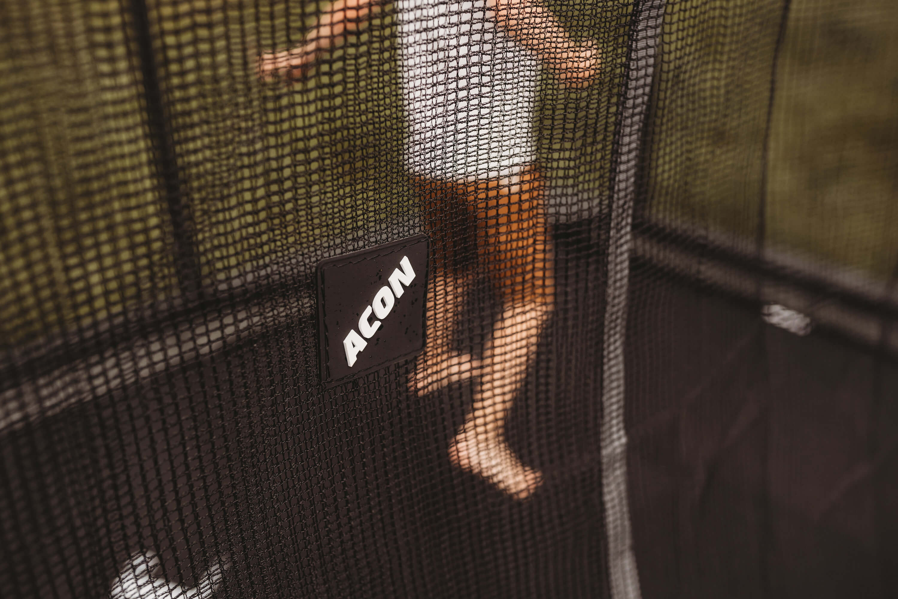 A kid jumping on an ACON trampoline with enclosure. The enclosure has a tightly-sewn mesh and a logo of the trampoline manufacturer Acon.