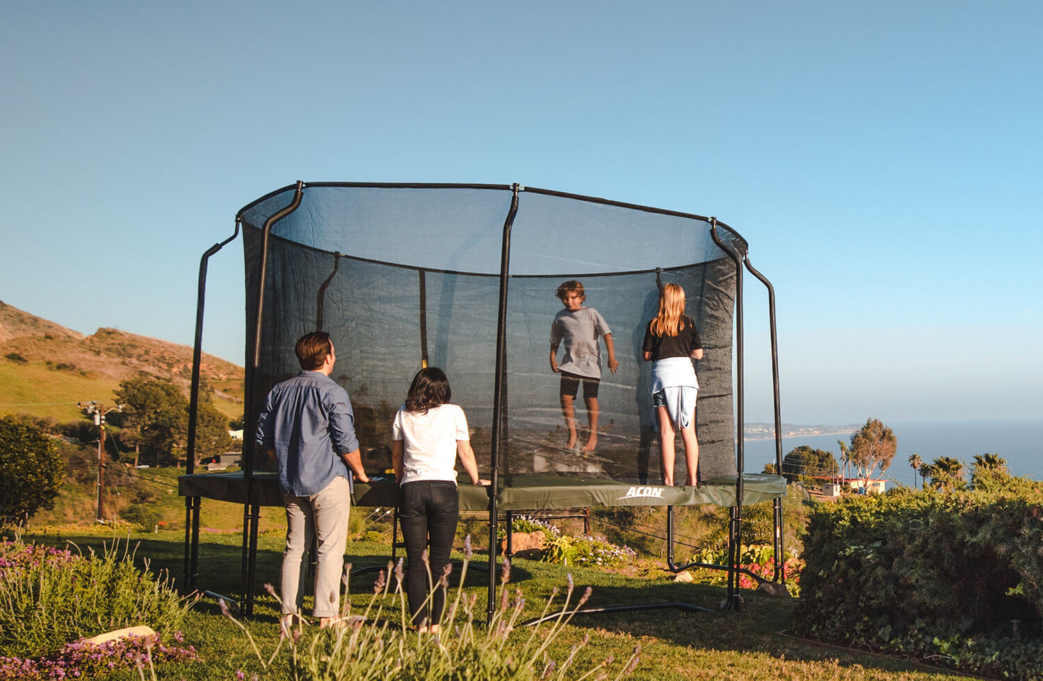 Boy juming on a trampoline with safety net. Dad, mom and sister watching.