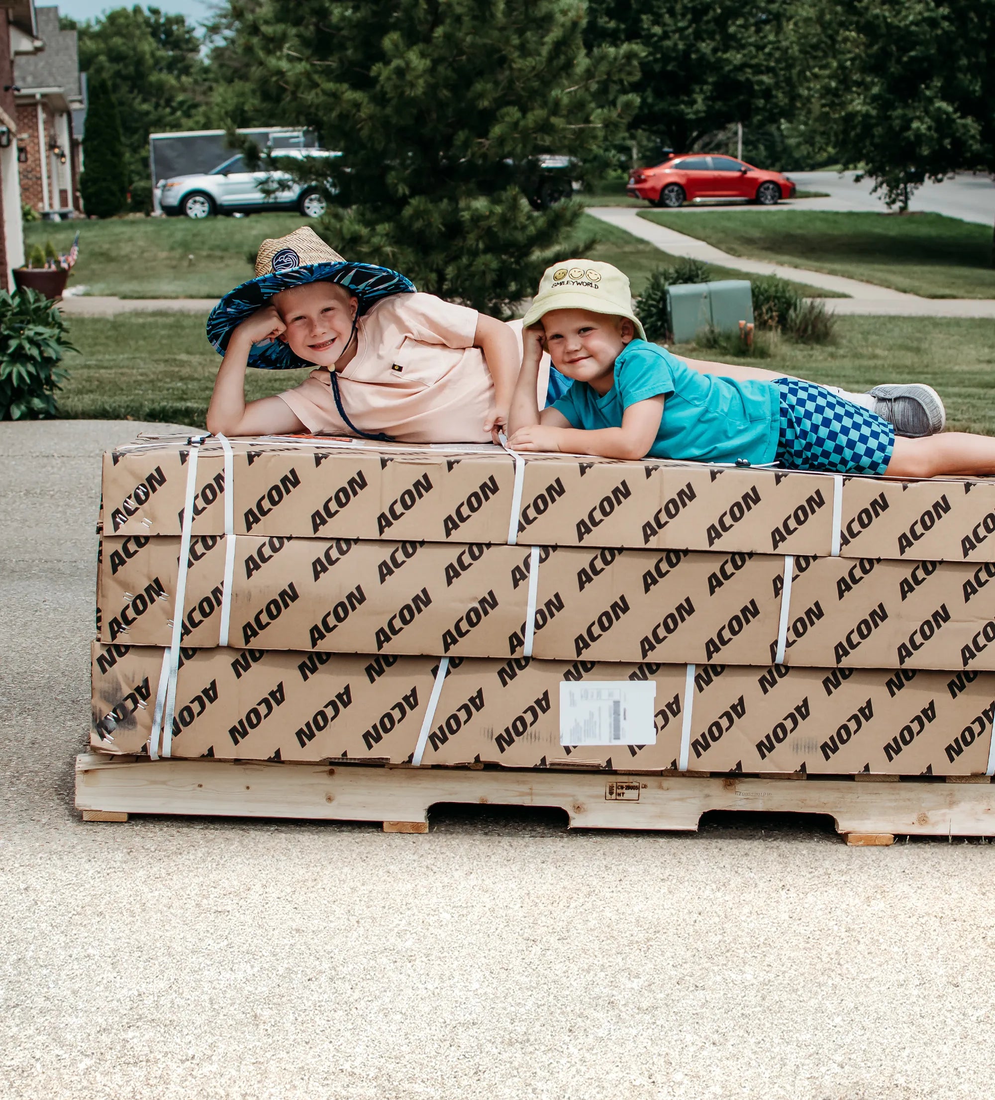 Two children laying on a pallet of trampoline cardboard boxes with 'ACON' branding, outdoors.