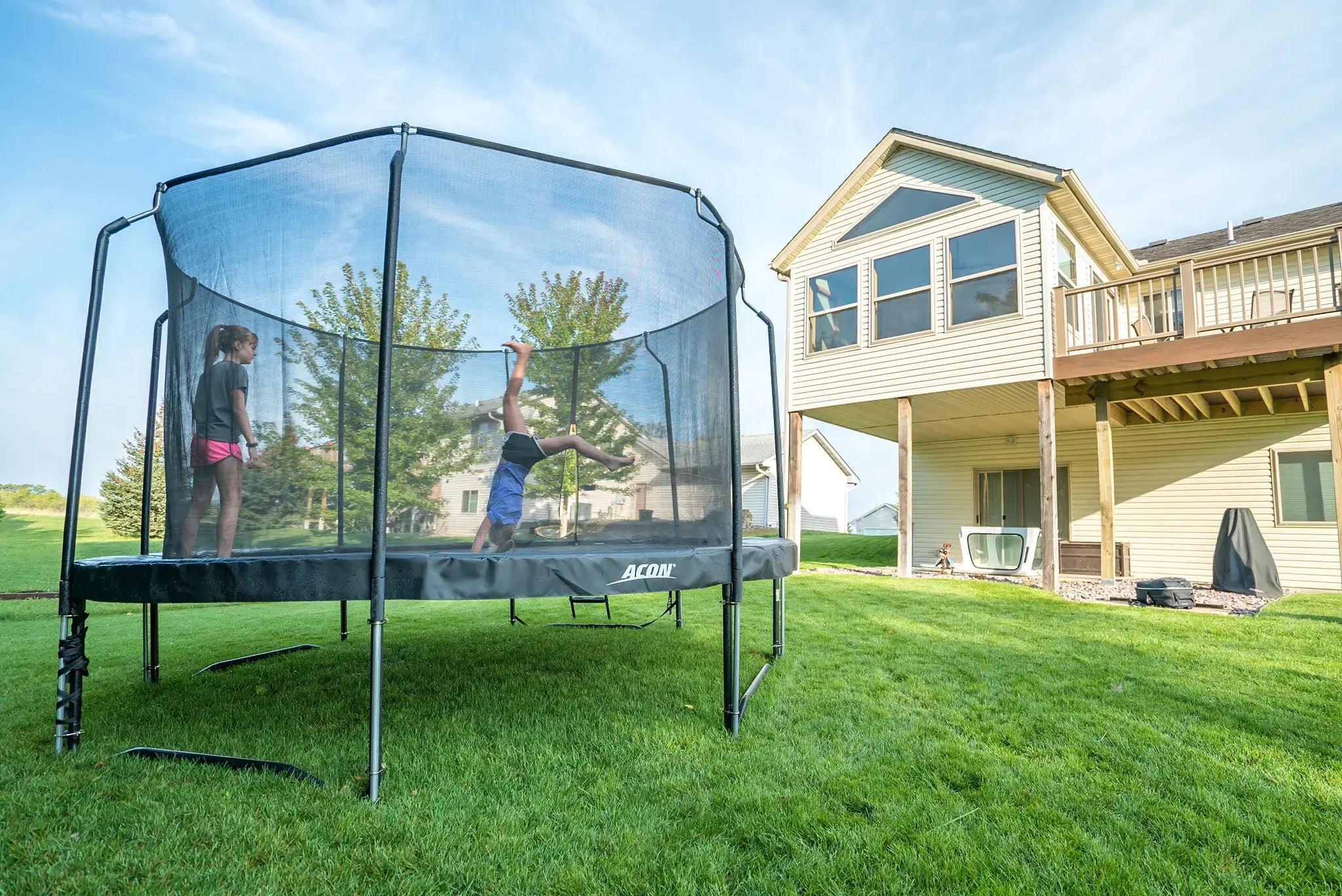 Two kids jumping on an Acon trampoline. One is doing a trick while the other is watching.