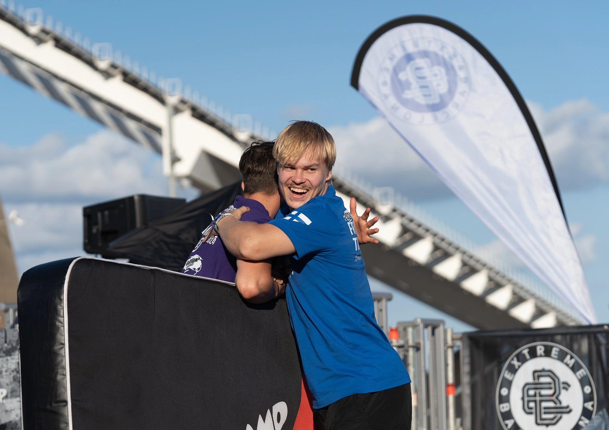 Two athletes hug near trampoline; one wears a blue Finland shirt, smiling after performance.