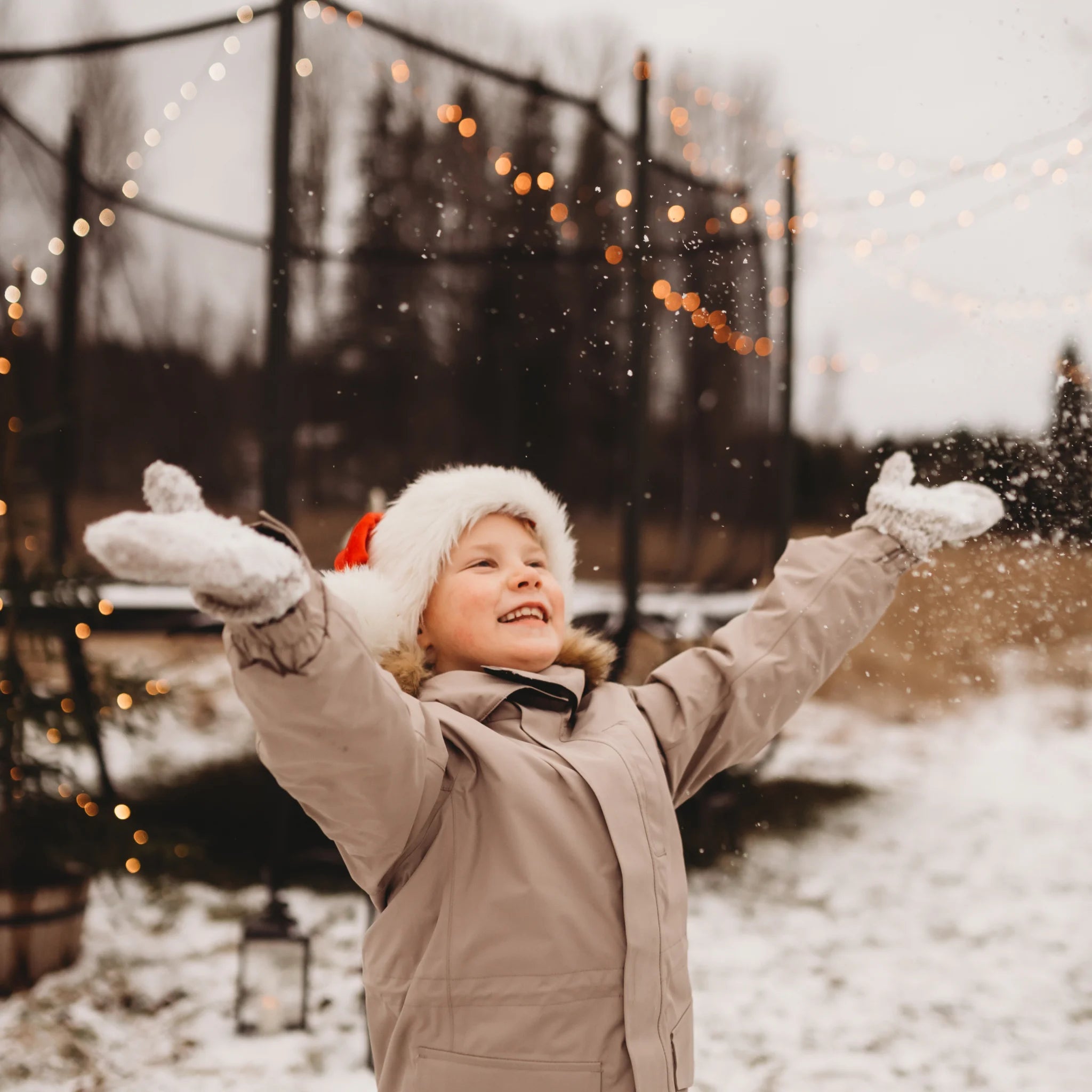 Happy child throwing snow in the air, with a trampoline decorated with Christmas lights in the beackground.