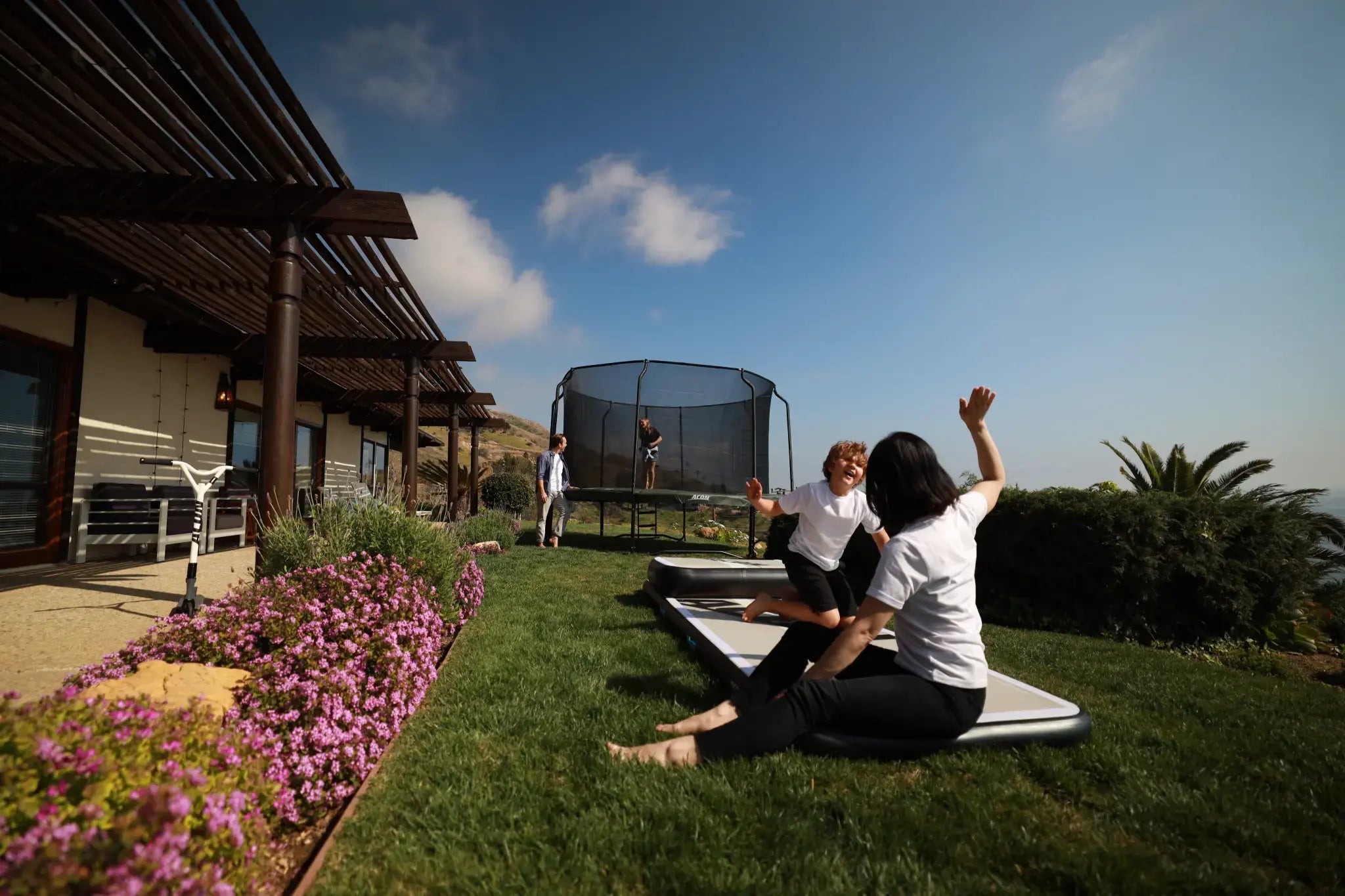 Woman high-fiving a child while sitting on an airtrack in a sunny backyard. There is a trampoline in the barckground.