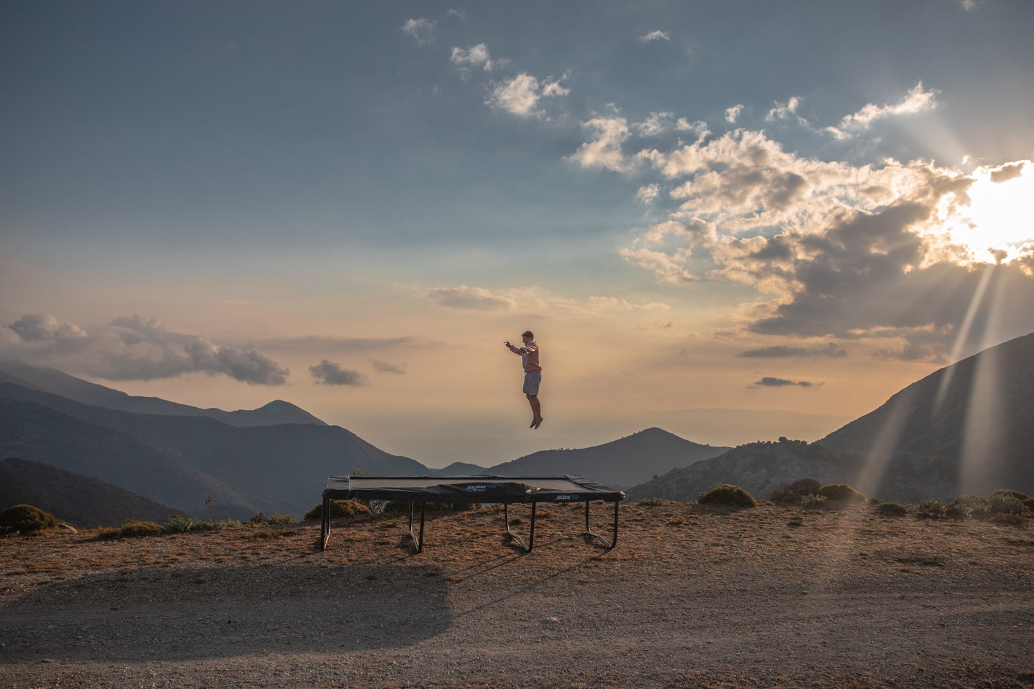 A guy jumping on an ACON trampoline outdoors with mountains and sunset as a background