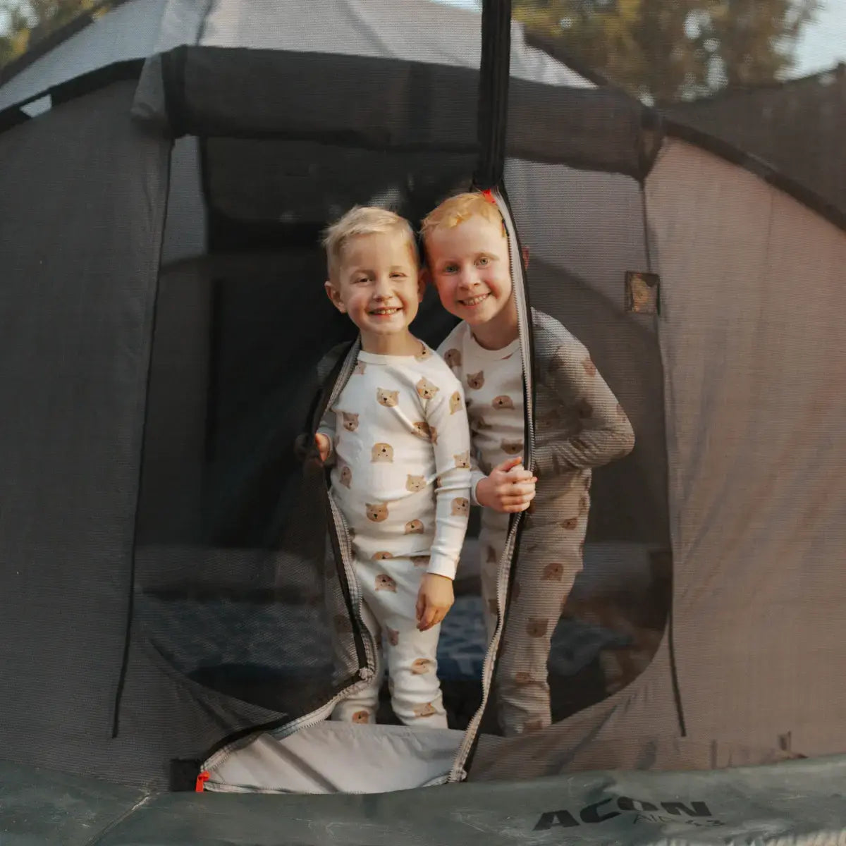 Two smiling kids in animal-print pajamas stand at the entrance of a trampoline tent, holding the zippered mesh door open.