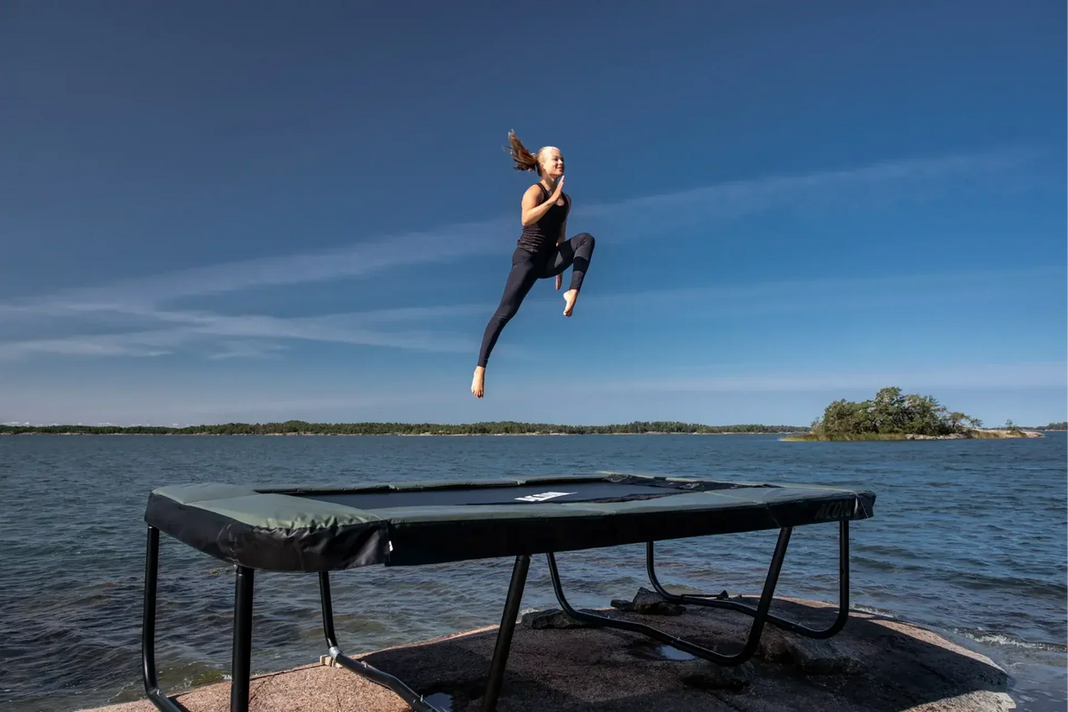 Woman jumping high on an Acon square garden trampoline with ocean on the background.