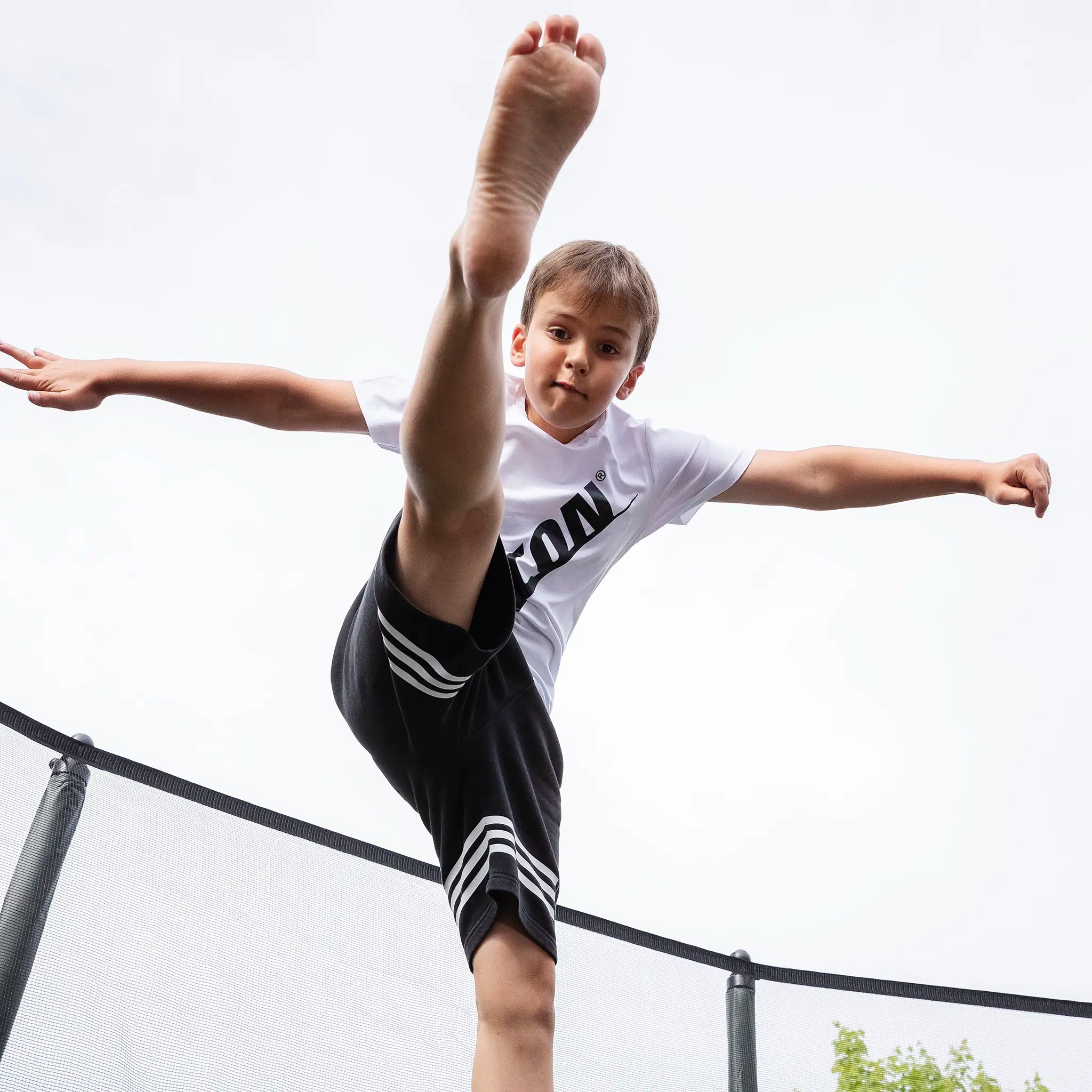 A boy performing a high kick on a trampoline, with one leg extended above his head. He is wearing a white Acon t-shirt. The background shows green trees and a cloudy sky.
