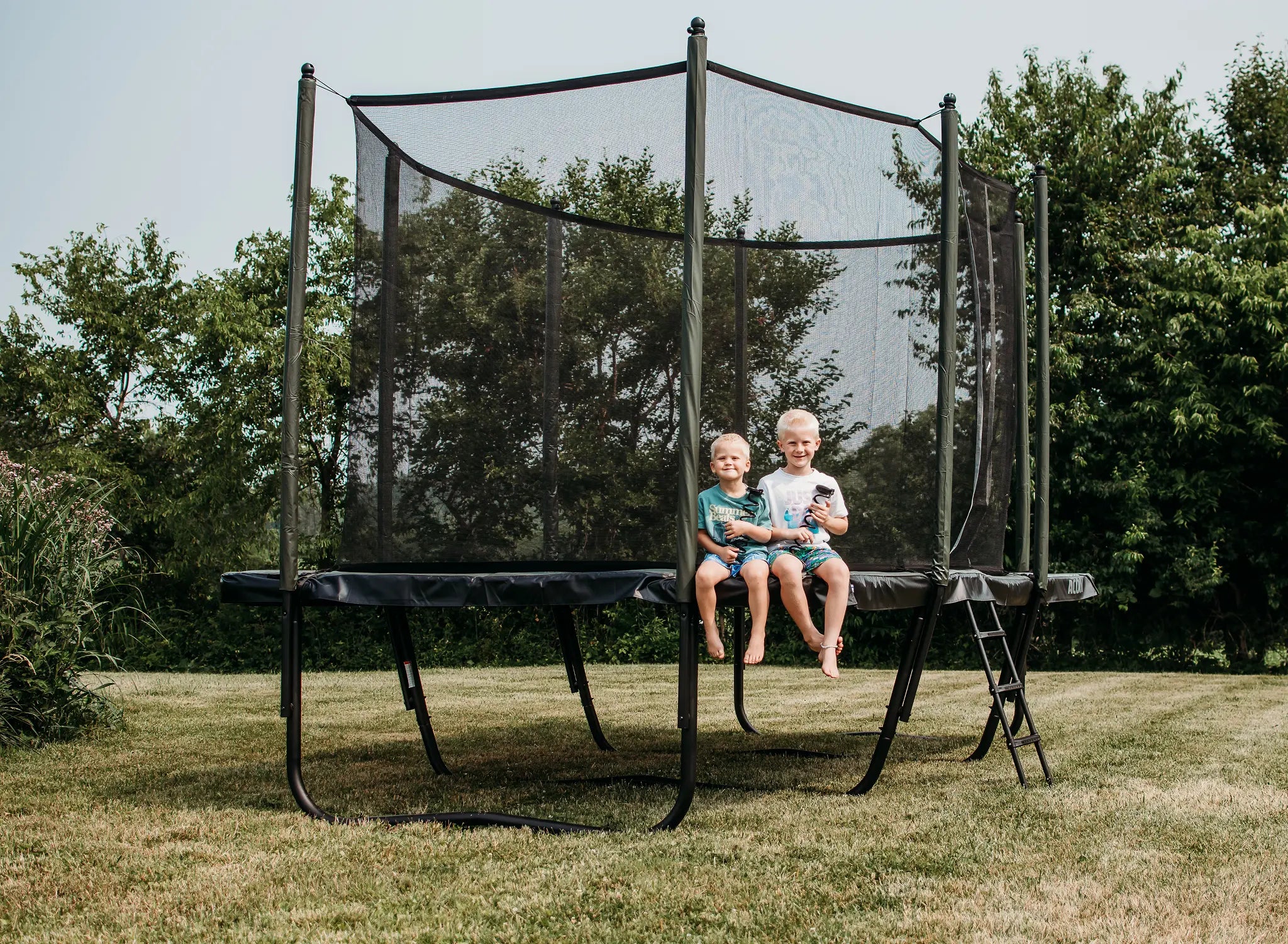 Kids sitting on a rectangular Acon trampoline with safety net in a backyard.