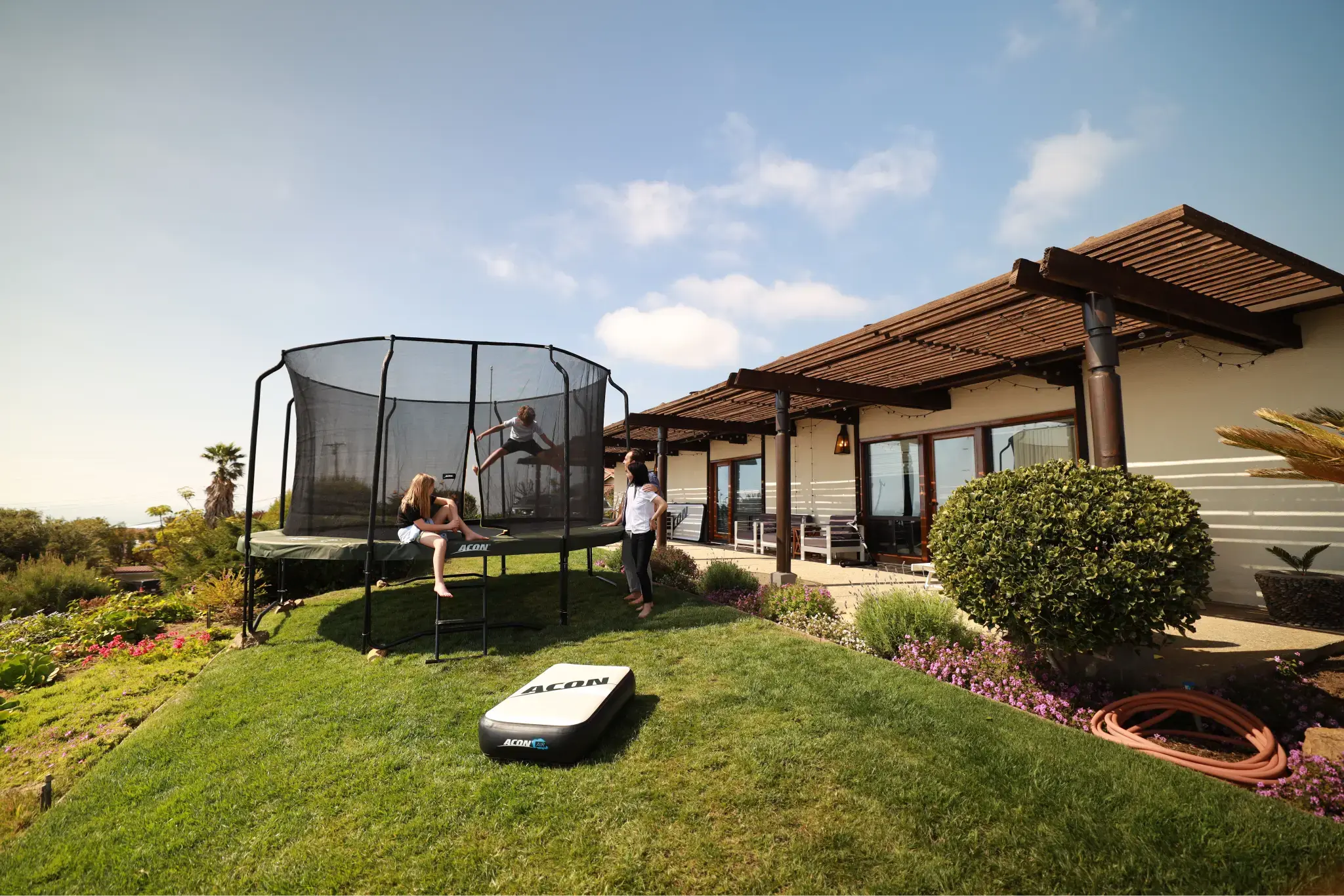 A family enjoys an Acon trampoline in a sunny backyard, with one child jumping inside the safety net while others watch and relax nearby.