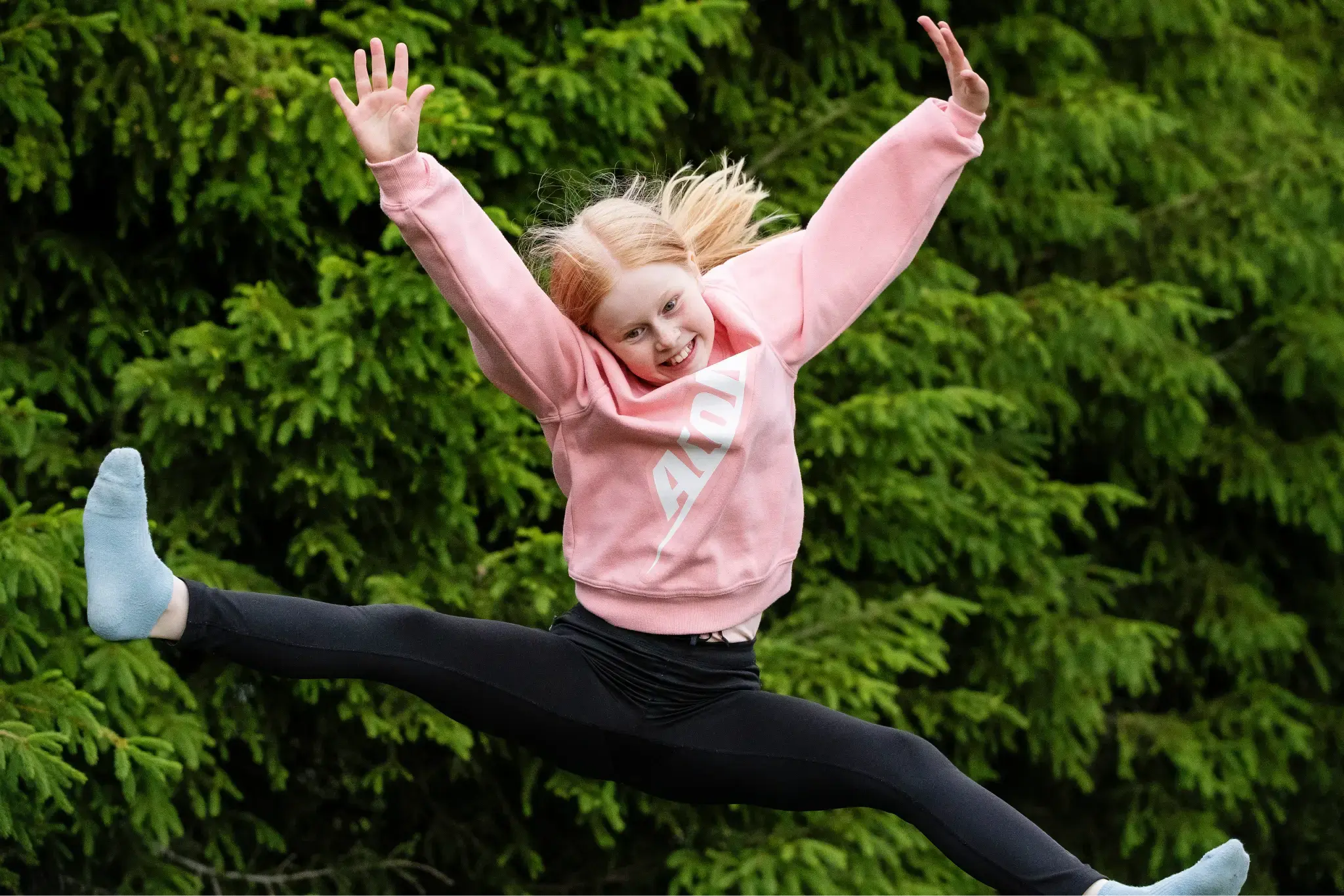 A young girl in a pink Acon hoodie and black leggings performs a split jump on a trampoline, smiling with arms raised against a backdrop of green pine trees.