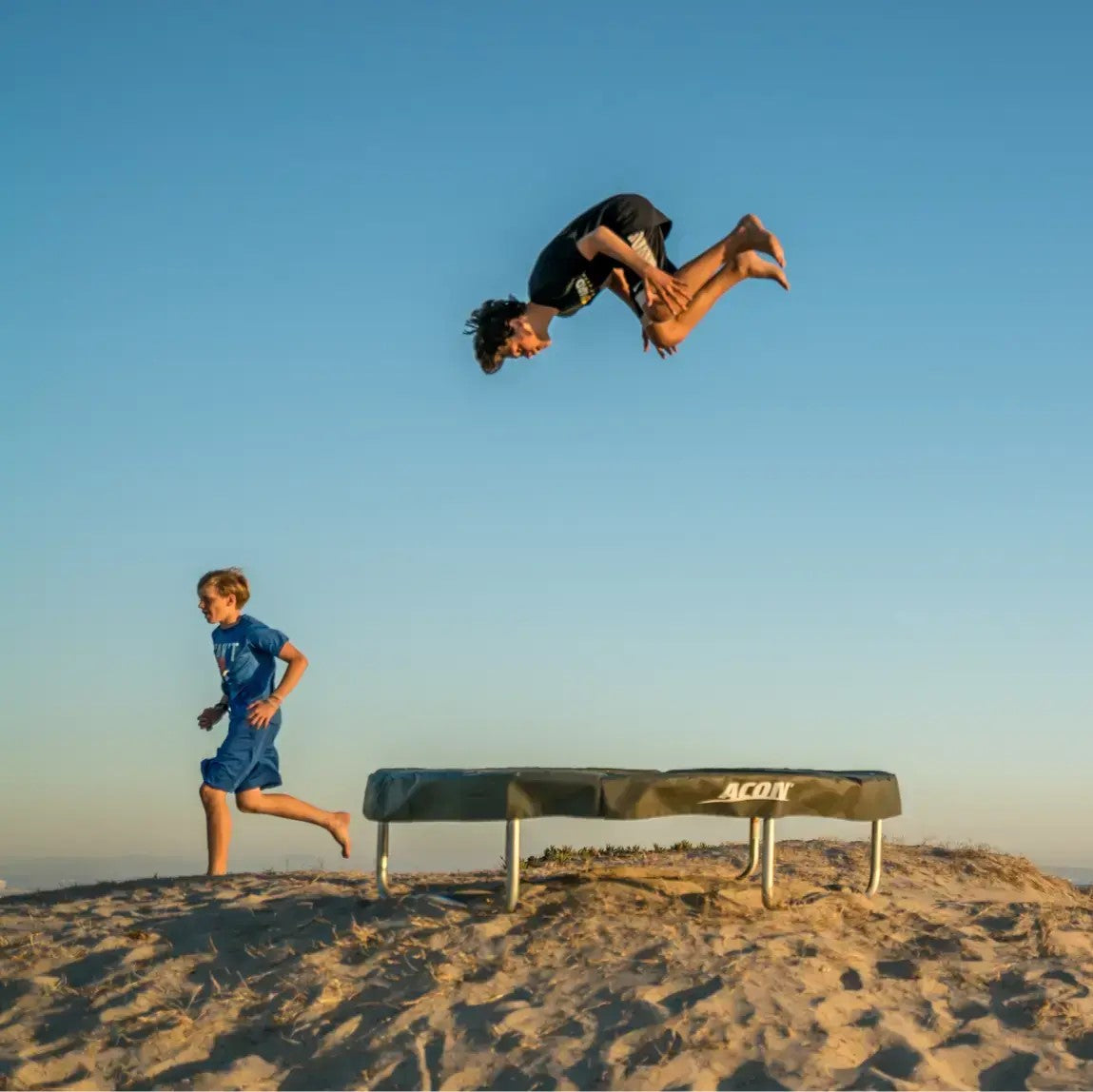 Two boys enjoying a trampoline on a sandy beach at sunset. One performs an acrobatic flip while the other runs nearby, with clear sky in the background.