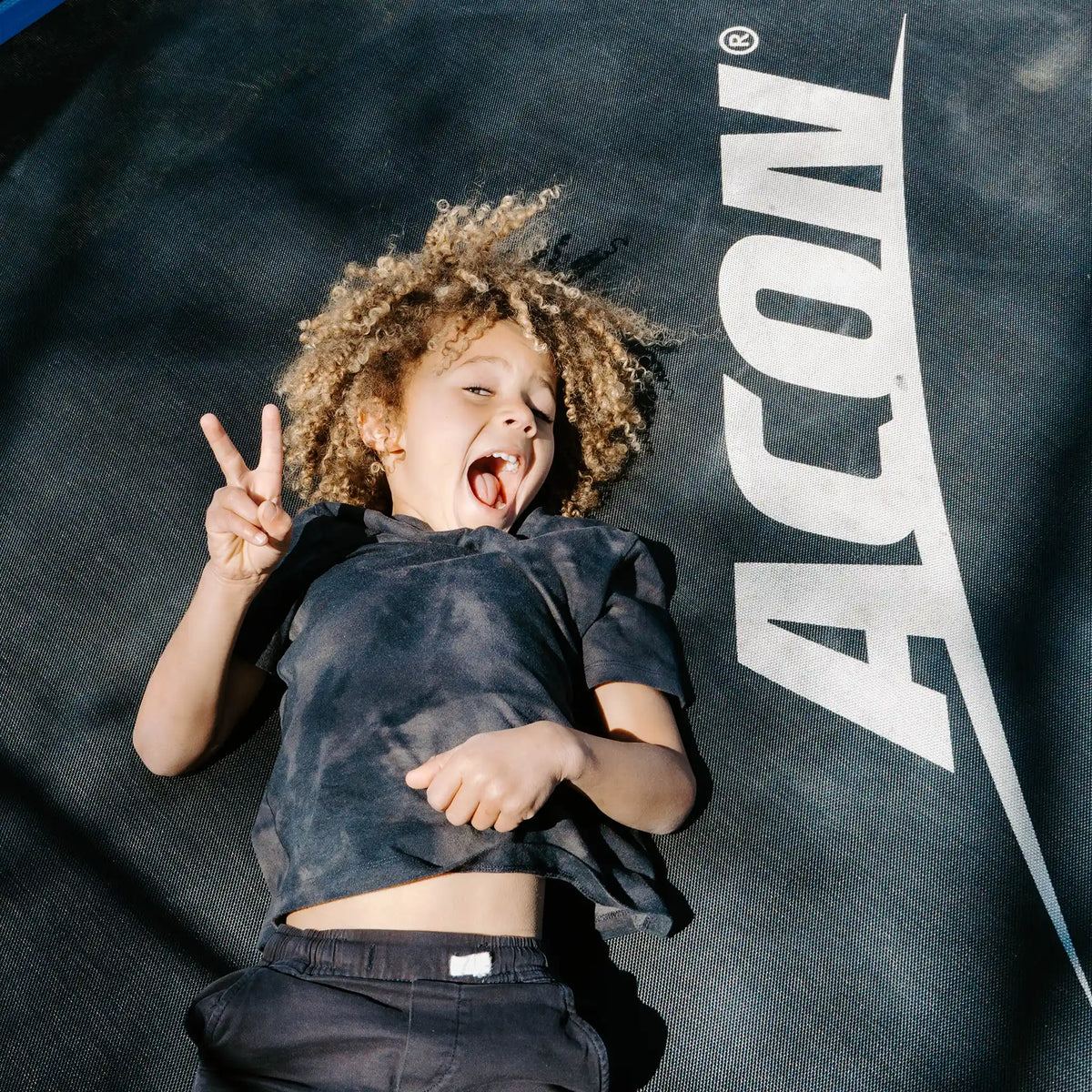 Young child laying on a Acon trampoline, showing peace sign and smiling.