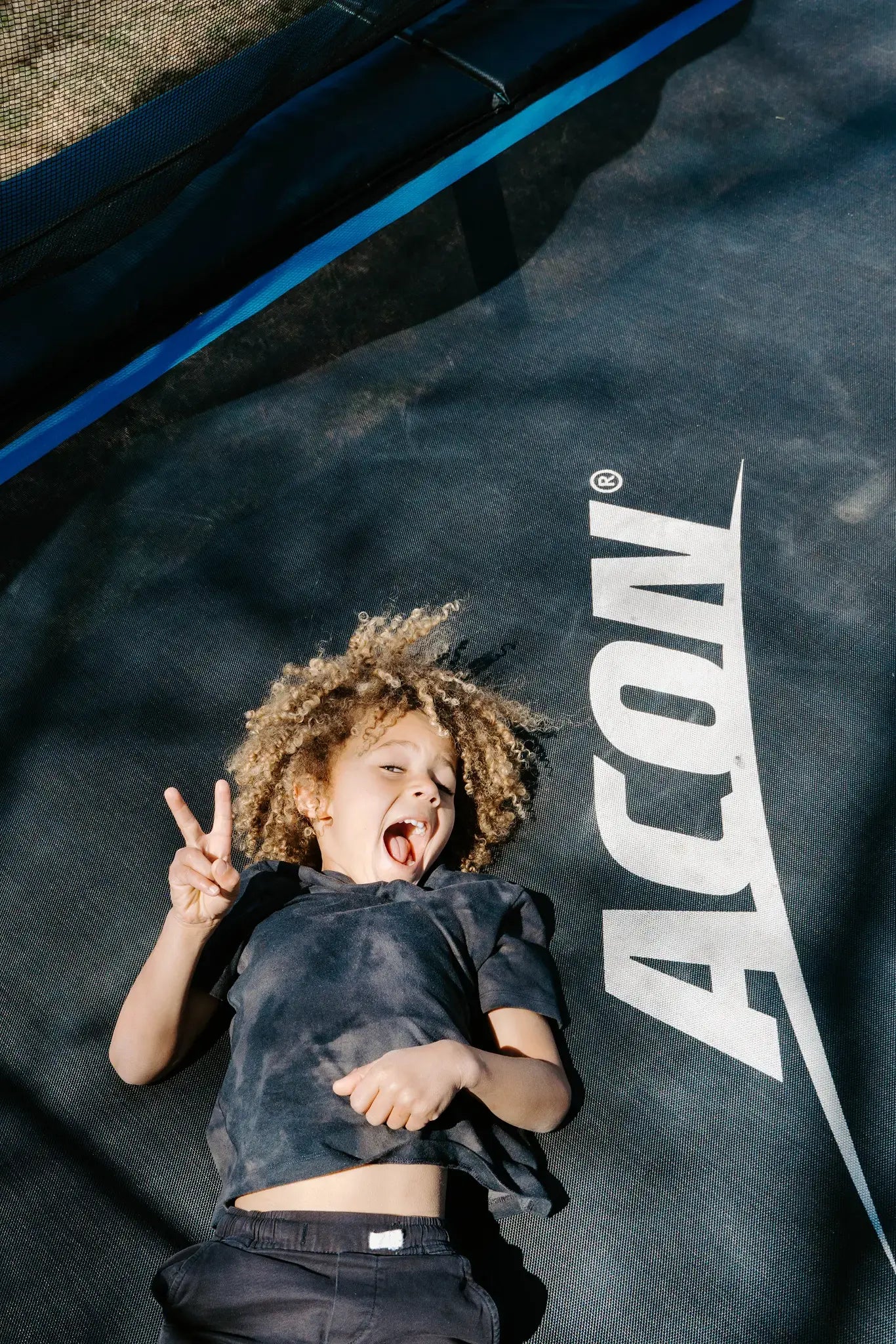 Young child laying on a Acon trampoline, showing peace sign and smiling. 