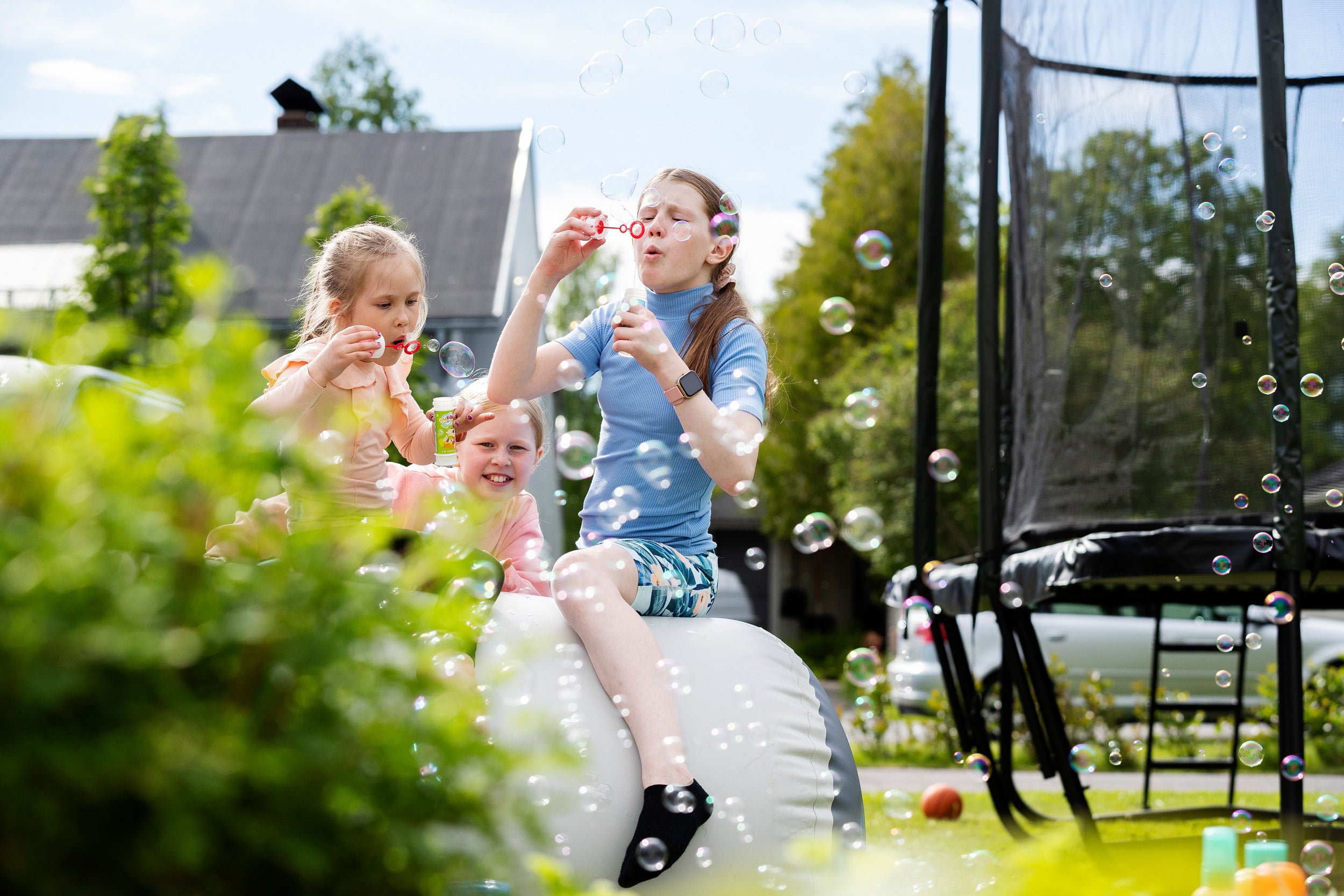 Kids blowing soap bubbles while sitting on an Airroll. There's a trampoline and green yard in the background.