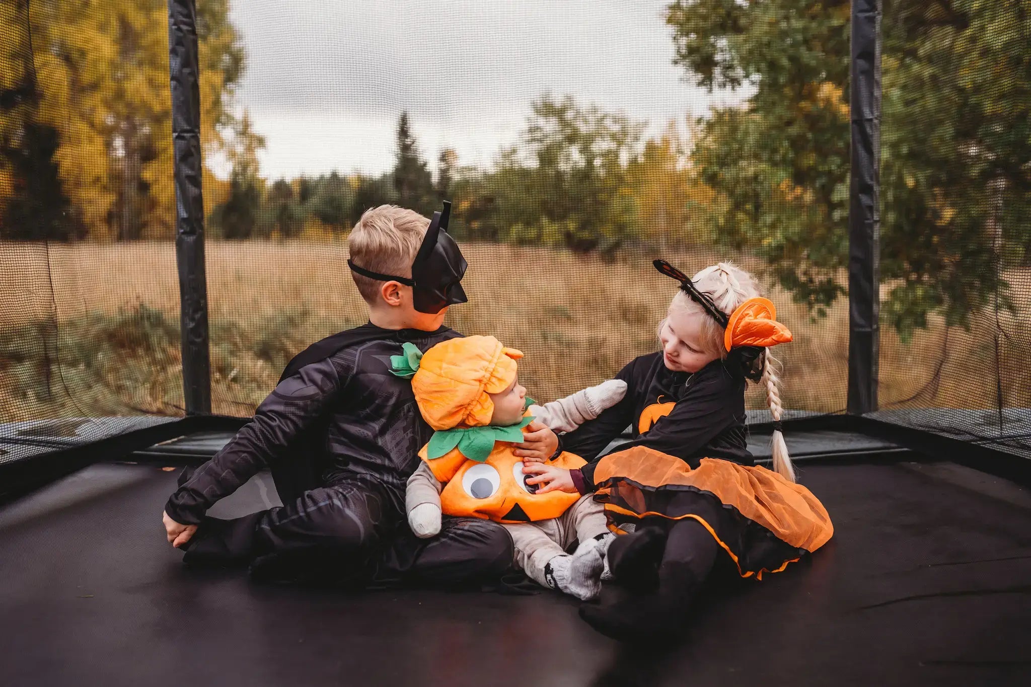 Girl, boy and a baby sitting on an Acon trampoline and wearing halloween costumes 