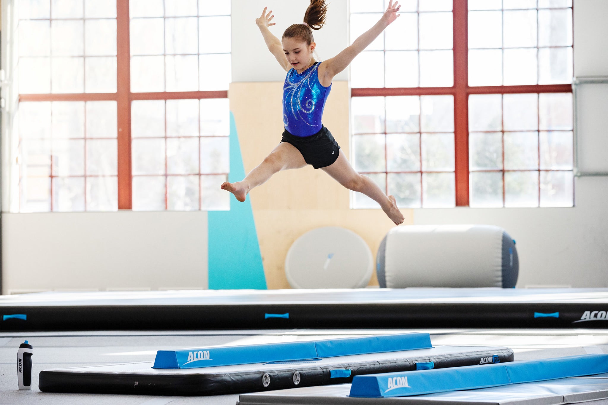 Girl doing a split leap on ACON gymnastics mat with balancing beam. There are ACON airtracks, airspot and airrolls in the background.