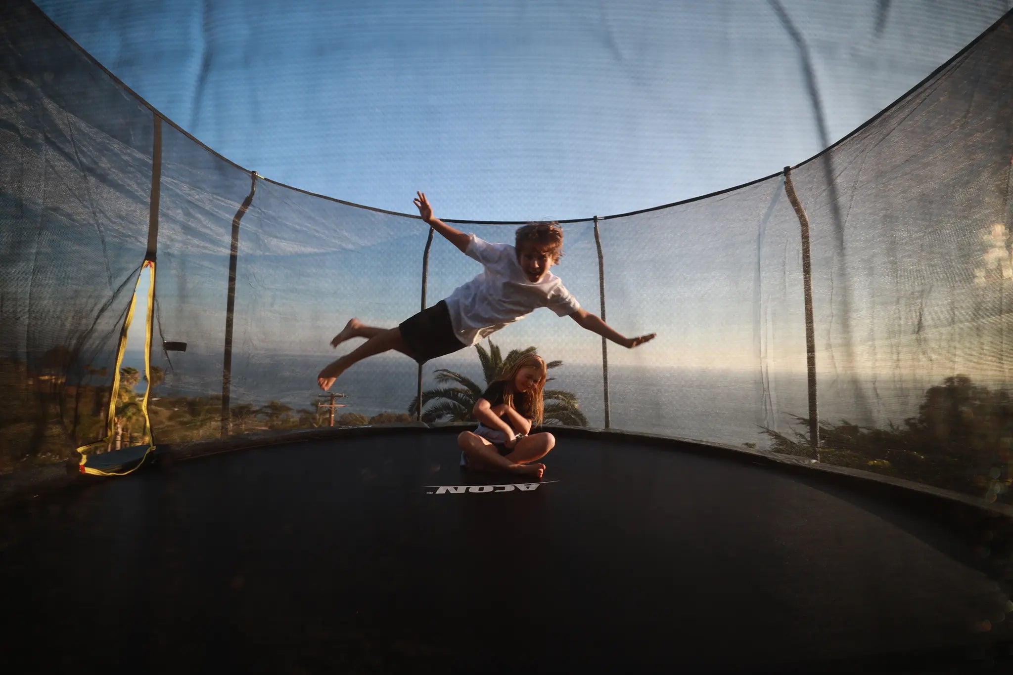 Two children playing on an Acon trampoline with a safety net, one jumping in the air while the other sits on the mat.