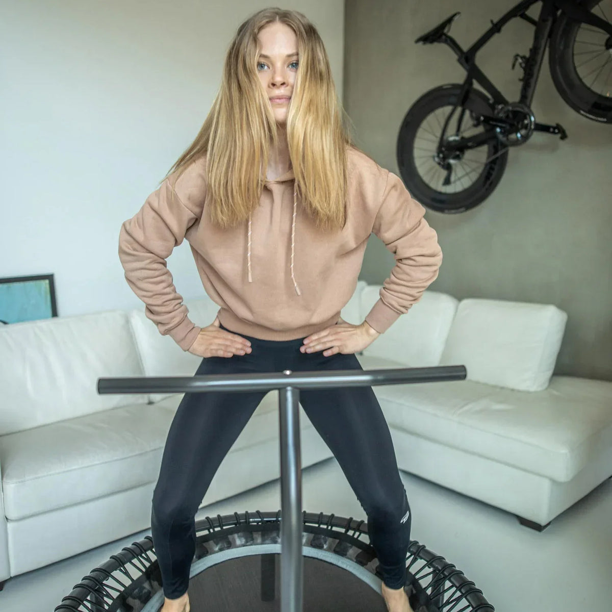 Woman standing on a rebounder with a handlebar in the livingroom. Behind her there's a white couch and a bike on the wall.