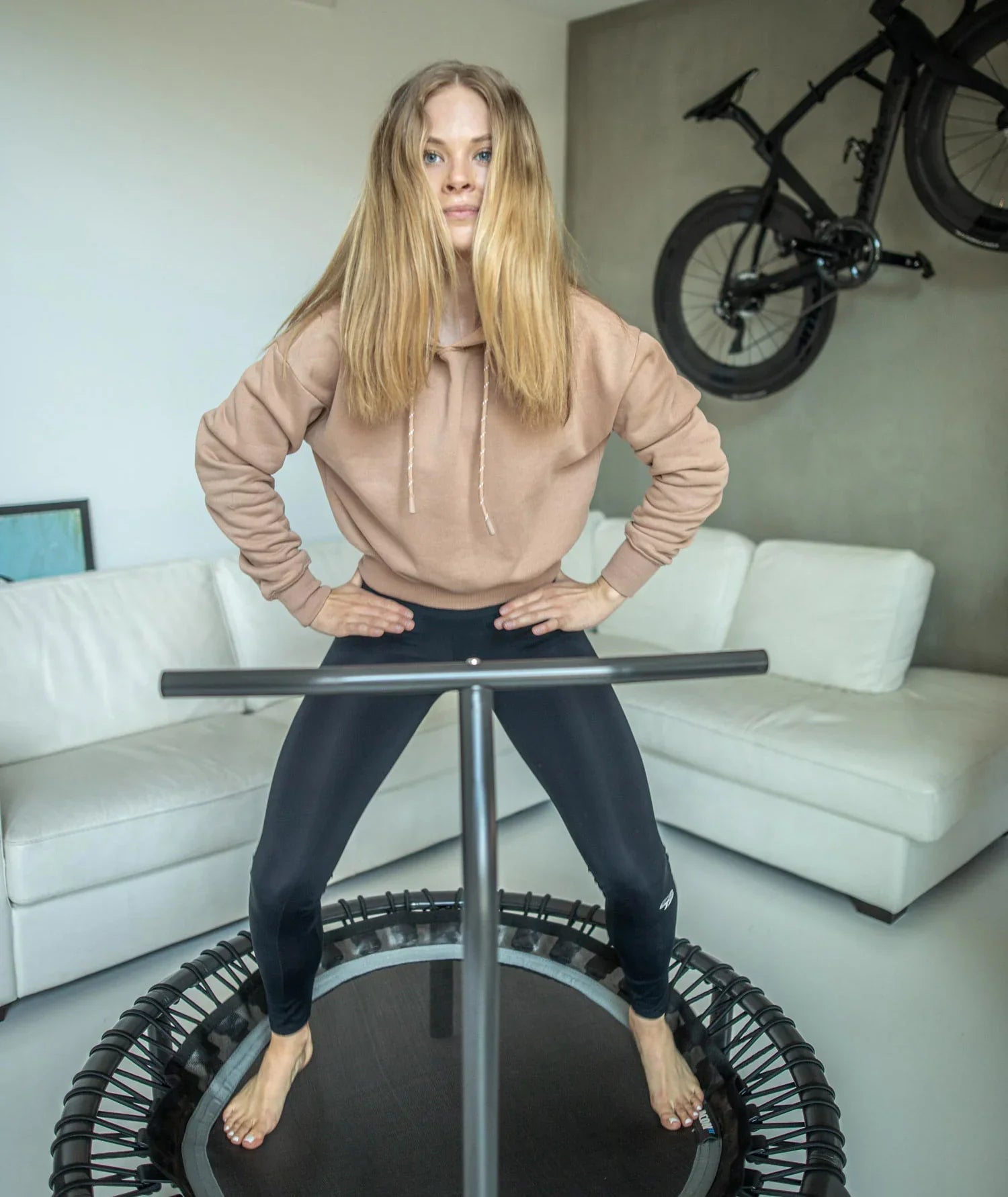 Woman standing on a rebounder with a handlebar in the livingroom. Behind her there's a white couch and a bike on the wall.