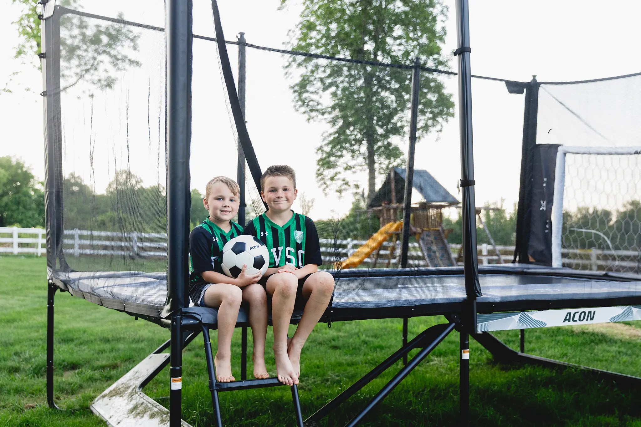 Kids sitting on an Acon X trampoline with safety net, holding a soccer ball in a backyard.