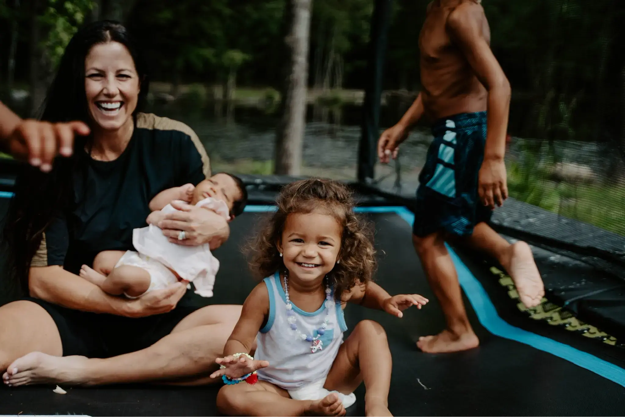 Smiling mother holding a baby while young children play and sit on a Acon trampoline, creating joyful family moments outdoors.