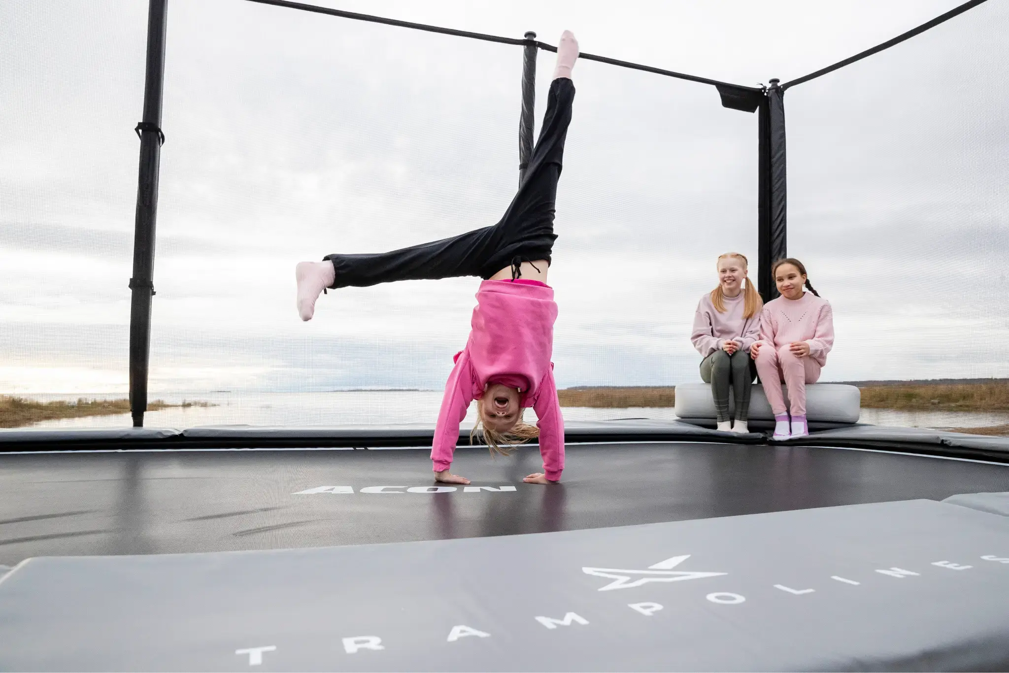 A child performing a handstand on a trampoline, with two other children sitting on the trampoline. The setting is outdoors by a calm body of water with grass in the background. The children are wearing casual, comfortable clothing. The trampoline is branded with the 'Acon' logo.