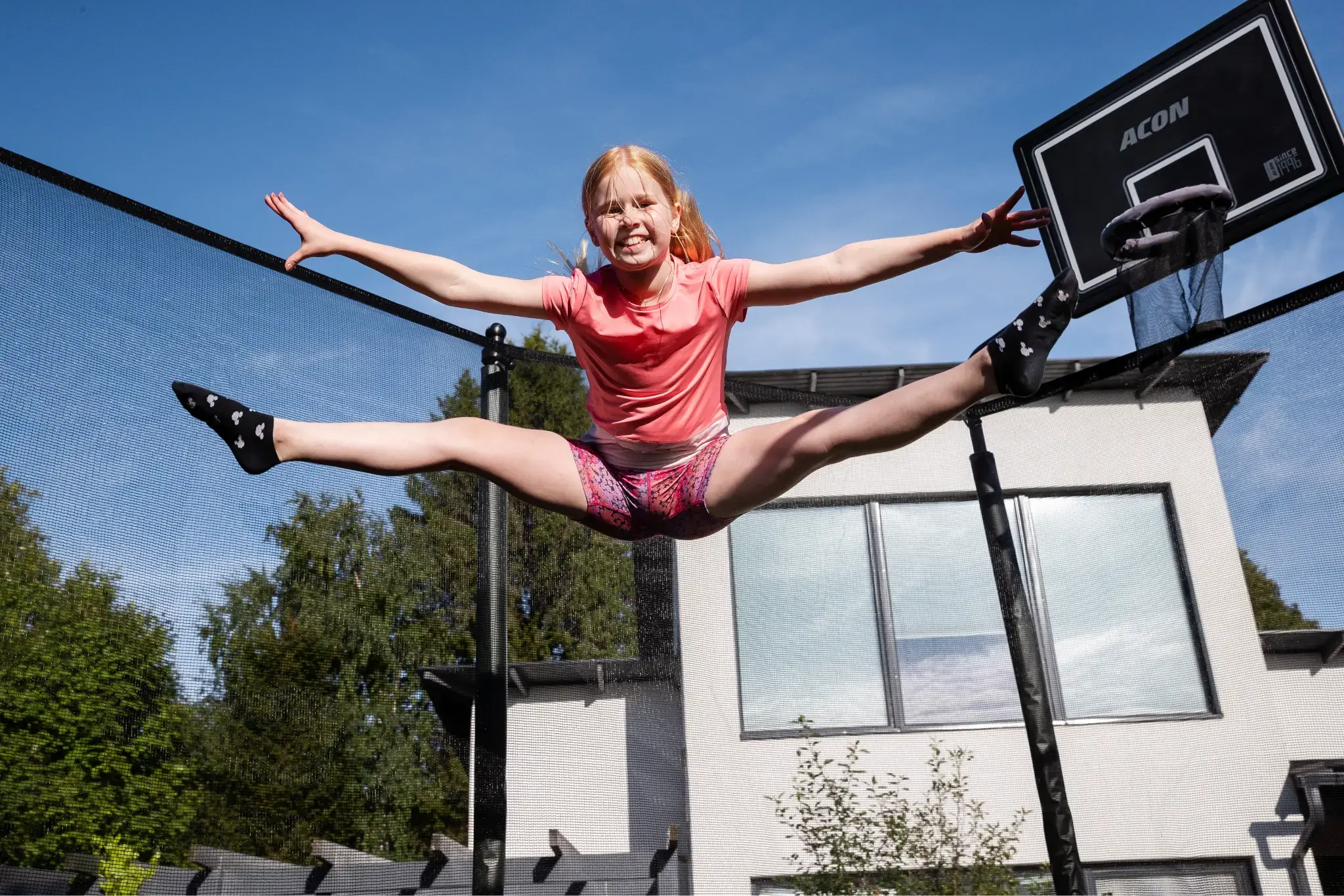 A girl performing a split mid-air on a trampoline, smiling and extending her arms and legs. She is wearing a pink t-shirt, patterned shorts, and black socks with white designs. The trampoline is equipped with a  Acon basketball hoop. A modern house is in the background, under a clear blue sky.