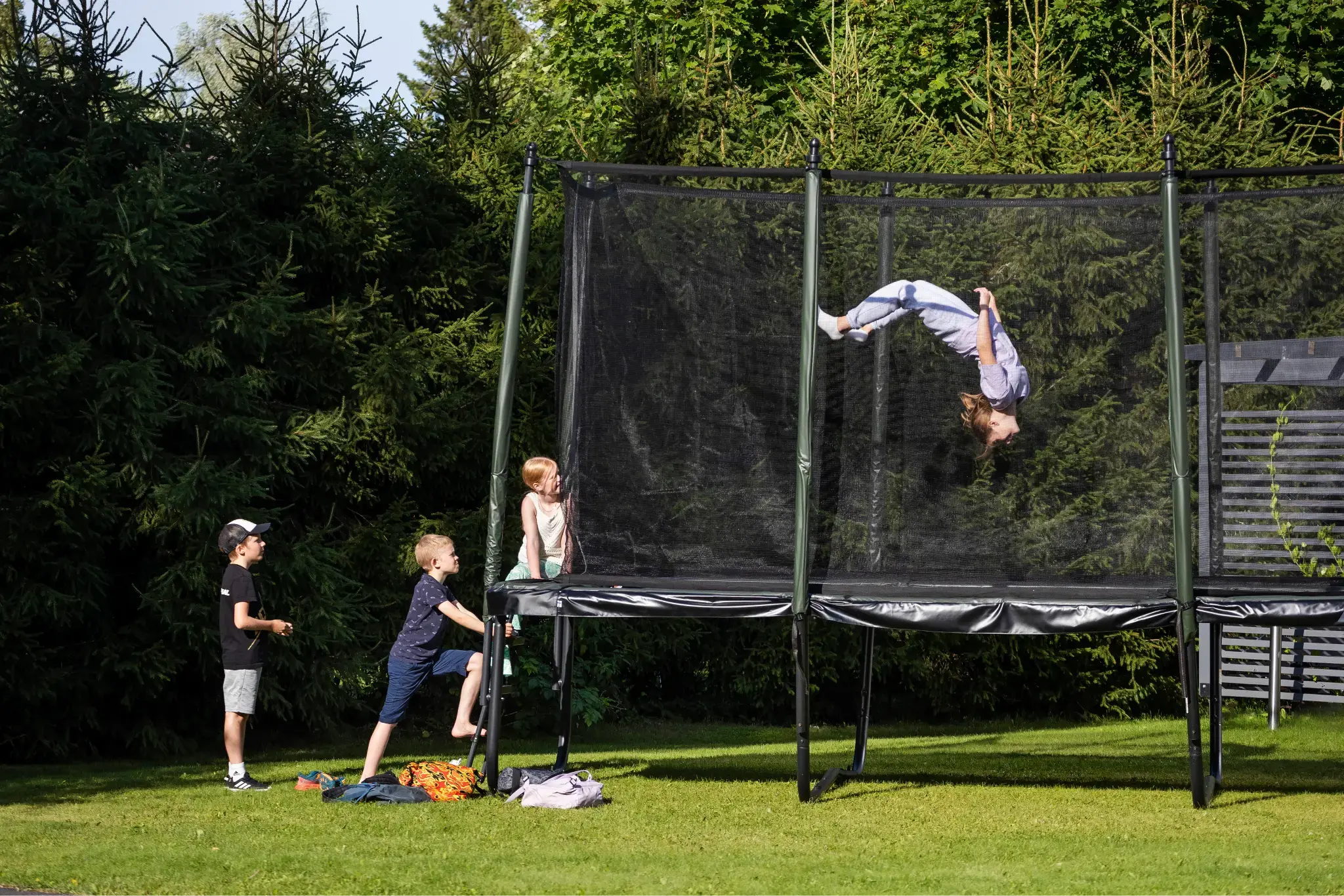 Children playing on a trampoline in a grassy outdoor setting. One child is performing a flip on the trampoline, while others are standing around, with one child climbing up the trampoline's side. The background features trees and a clear sky.