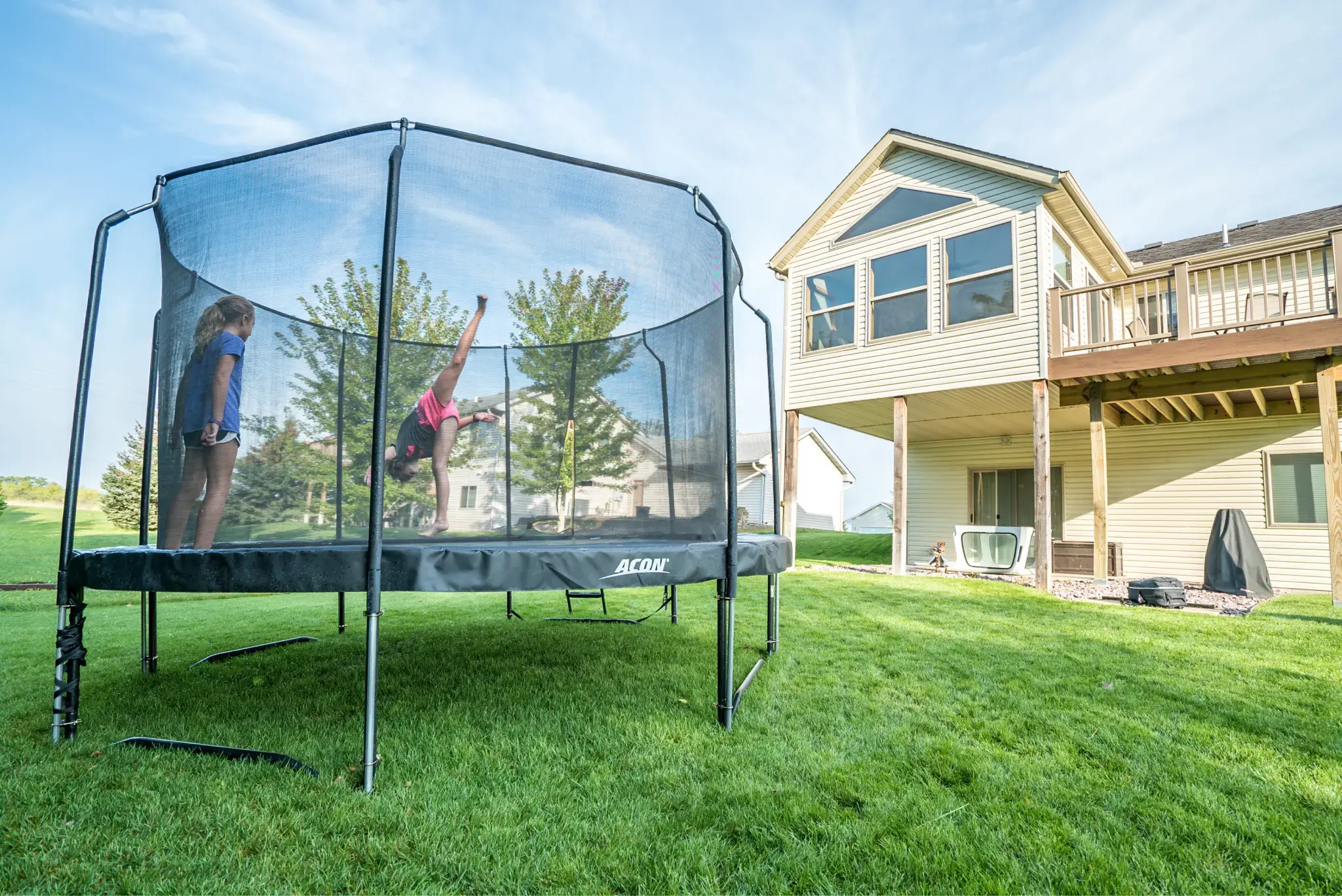 Two children playing on a large Acon trampoline with a safety net in a backyard, surrounded by green grass and a modern house in the background.