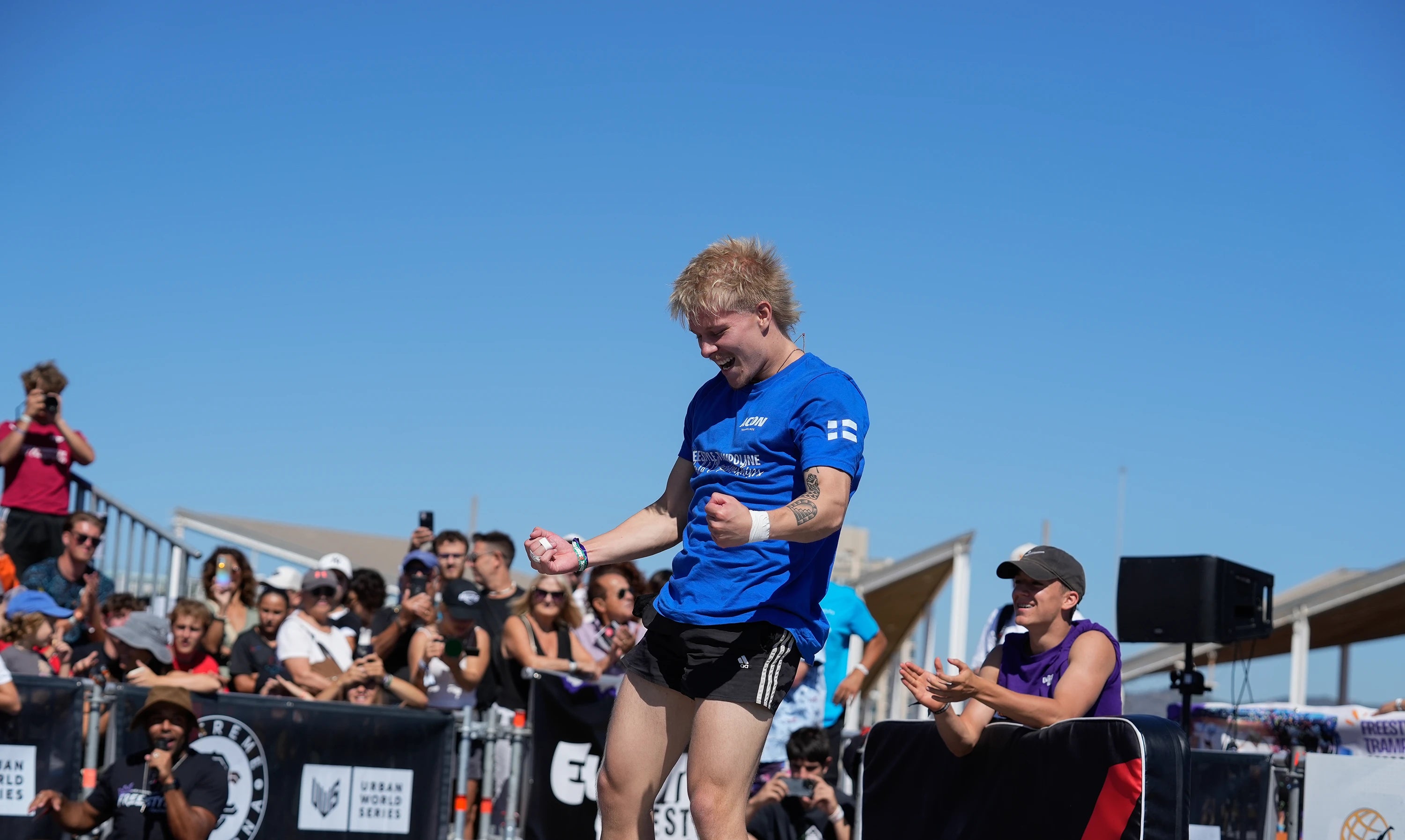 Man in blue shirt and black shorts on a trampoline with spectators in the background.