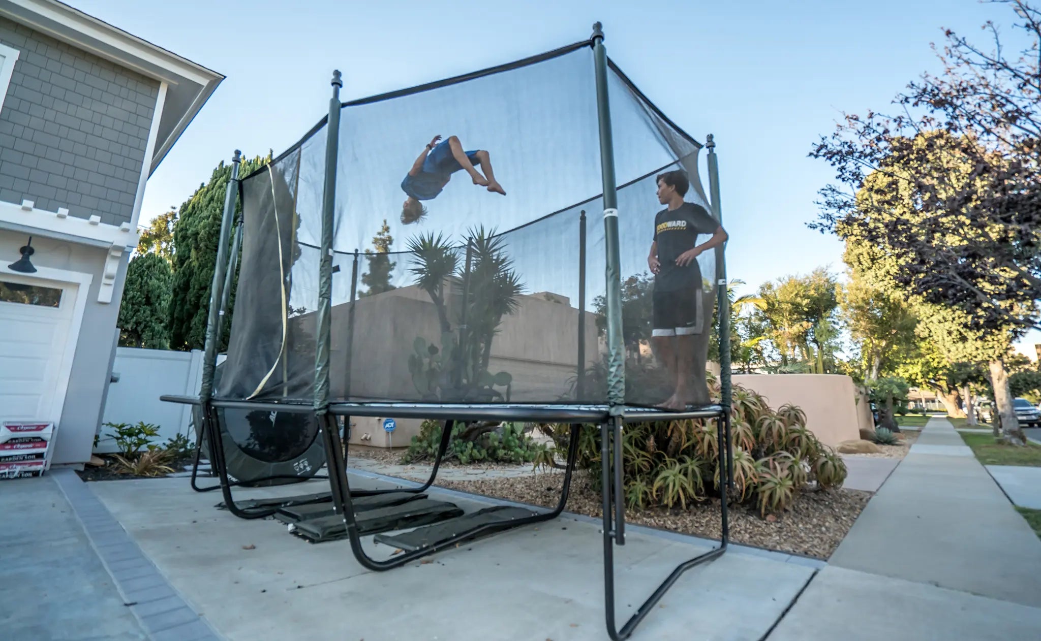 Two boys on an Acon trampoline with a safety net—one standing and one mid-flip.