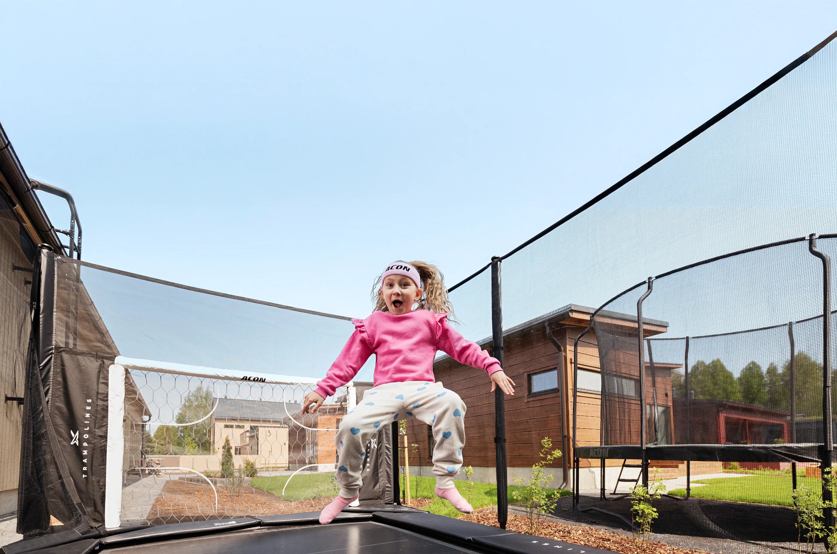 A joyful young girl in a pink shirt and Acon headband jumps on an Acon X Trampoline, her happy expression showing pure fun. Houses and blue sky behind her.