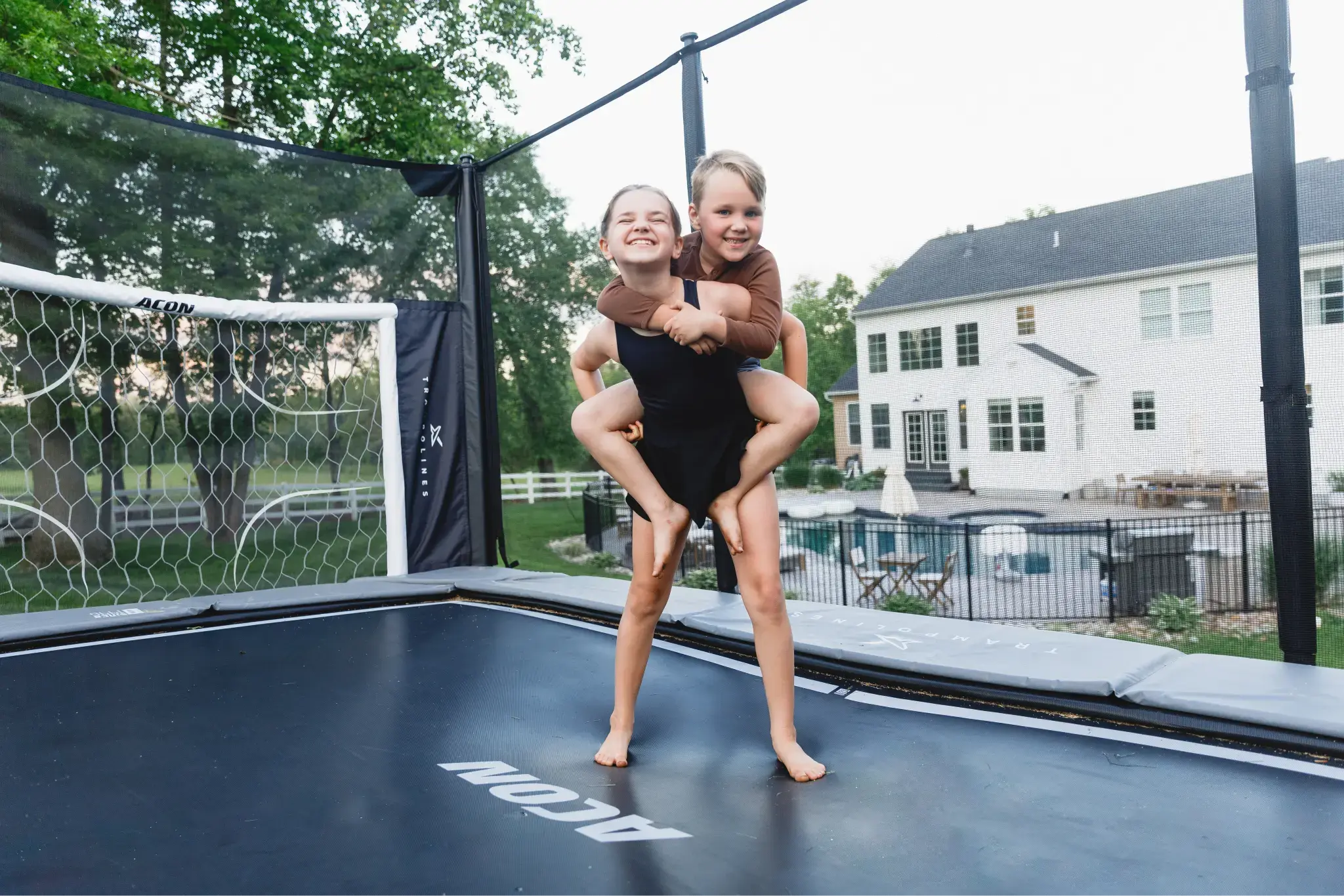 Two smiling kids on a trampoline in the backyard of a big house, one giving the other a piggyback ride in.