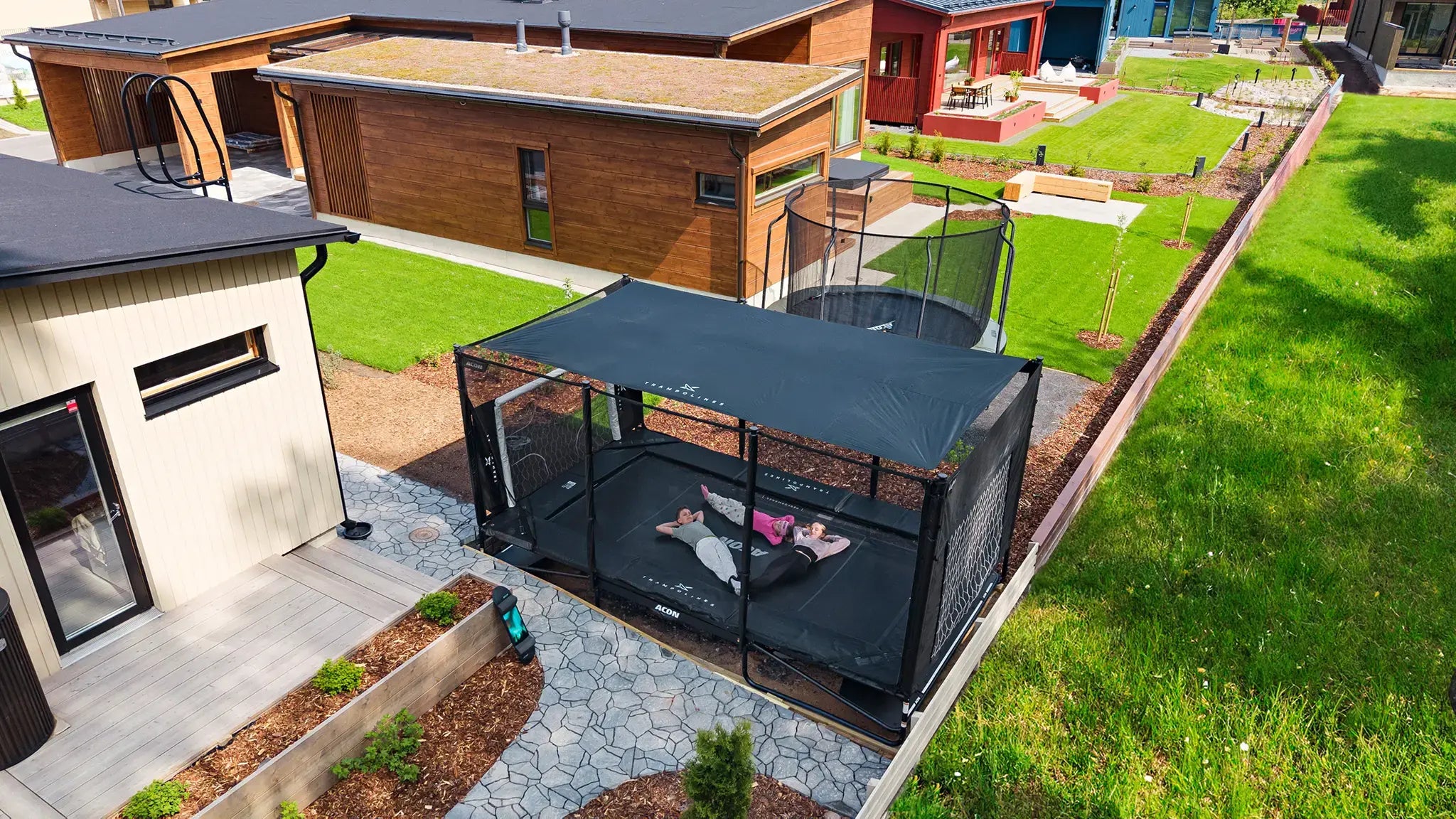 Three kids relaxing on an Acon X trampoline with a sun shade in a modern backyard.