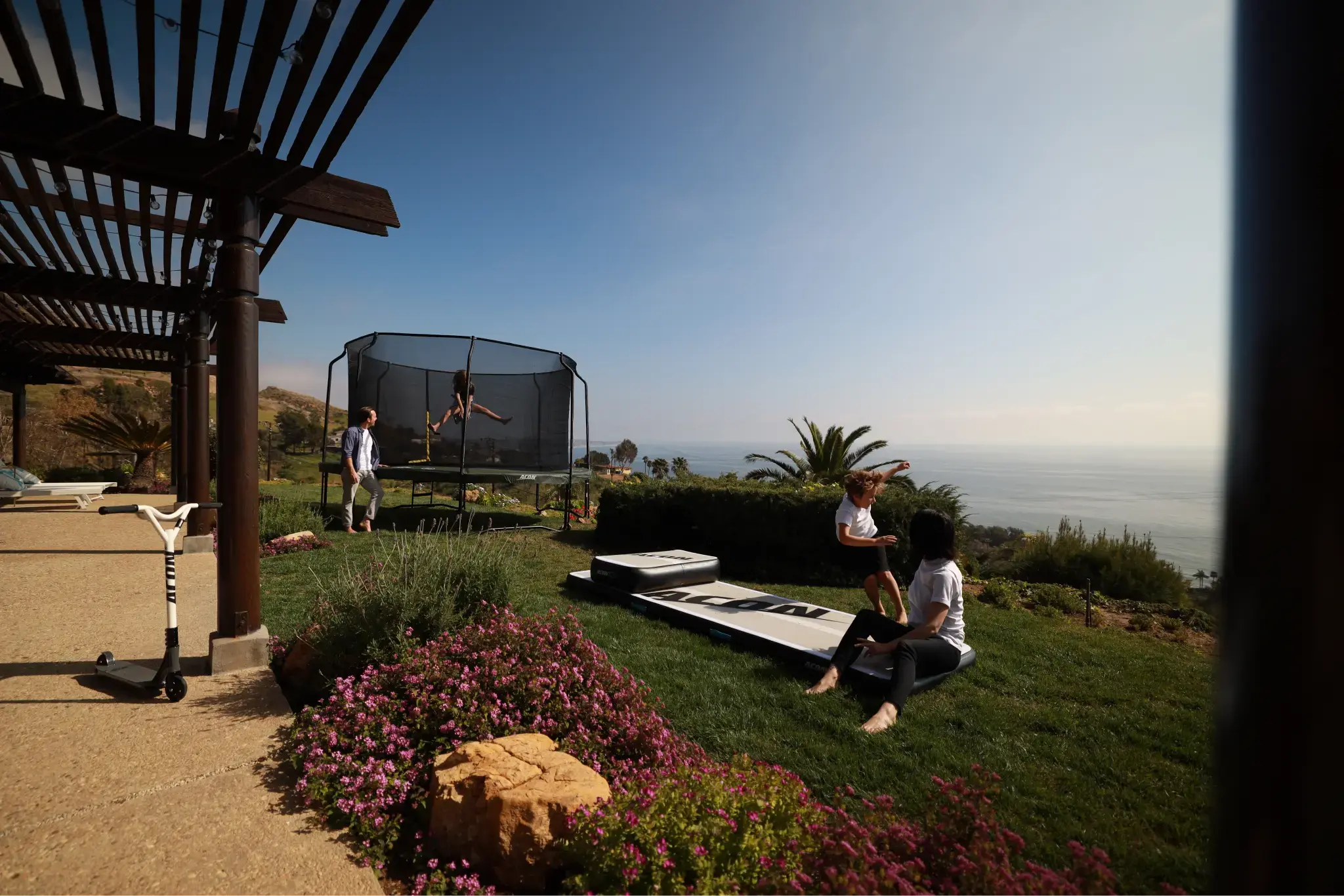 Family enjoying a backyard with a trampoline, an airtrack, and sea view, kids jumping and playing outside.