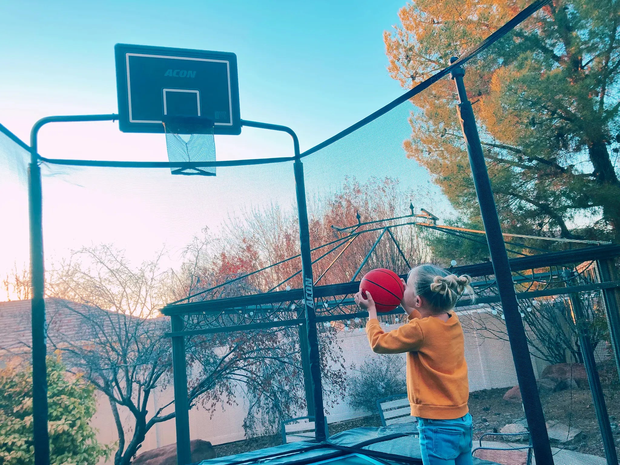 A little girl attempting to throw a basketball through a hoop on an Acon trampoline with net.