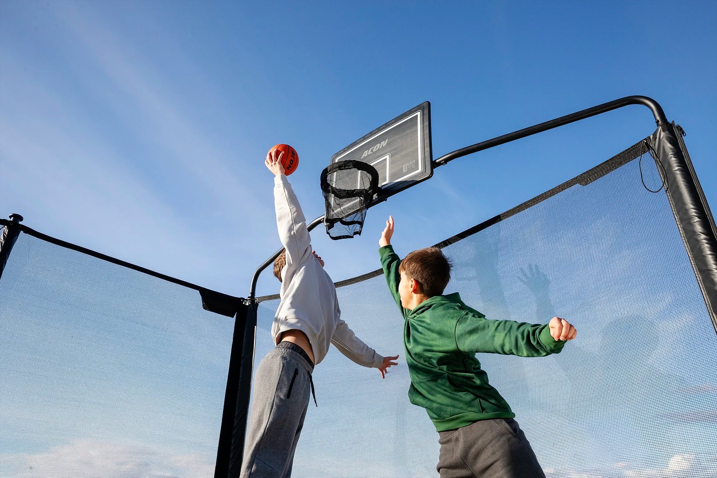 Two people playing basketball on an Acon X Trampoline outdoor with a clear blue sky.
