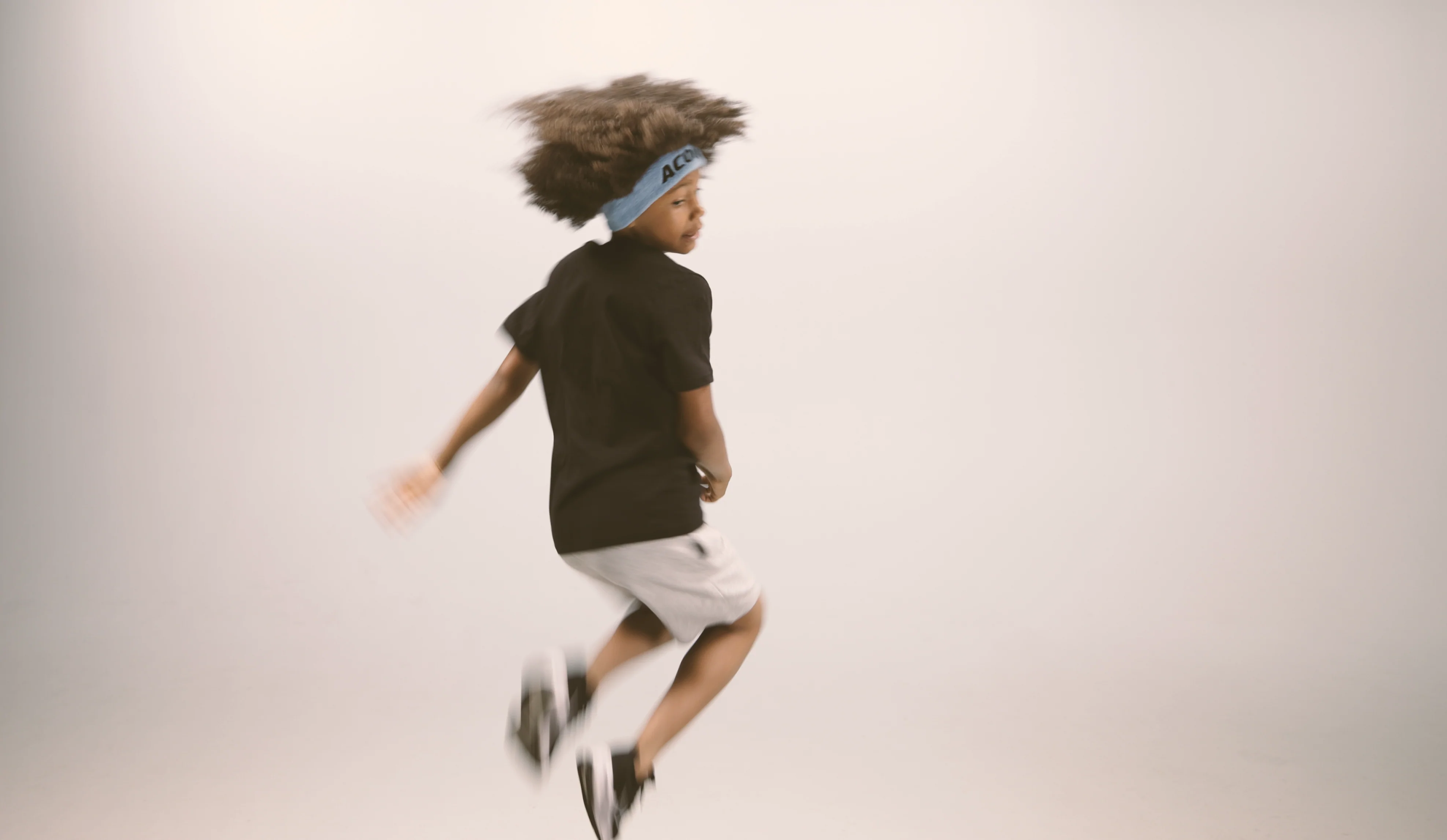 A jumping kid wearing an Acon headband, black t-shirt, white shorts and sneakers. Image is taken against beige background. 