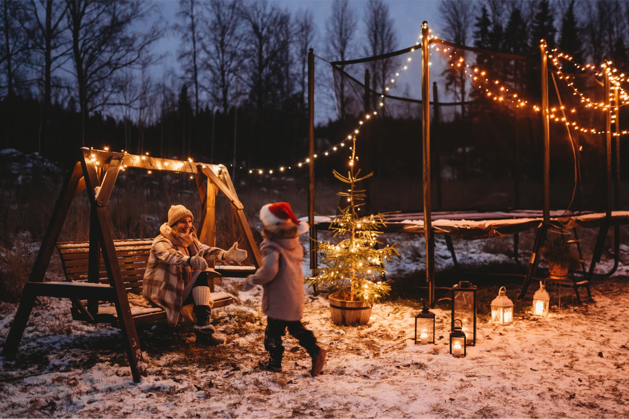 Child with a santa hat going to their mother on a swing. There's snow on the ground, and an Acon trampoline with Christmas lighting on the background.