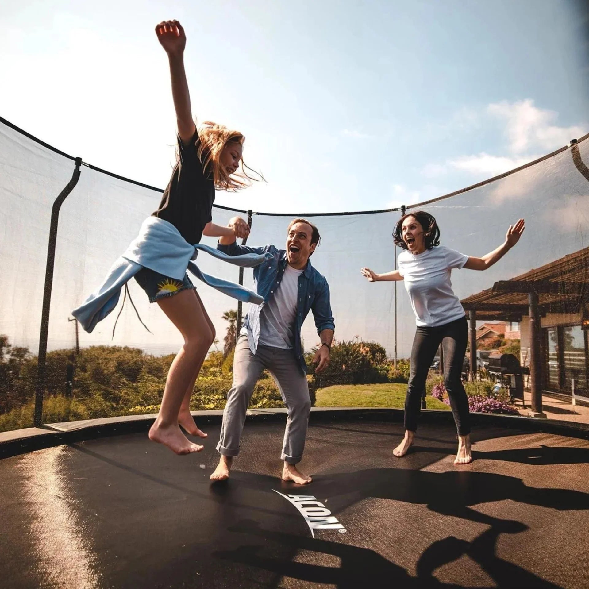 Parents and daughter are jumping and laughing on a round Acon trampoline with safety net. 