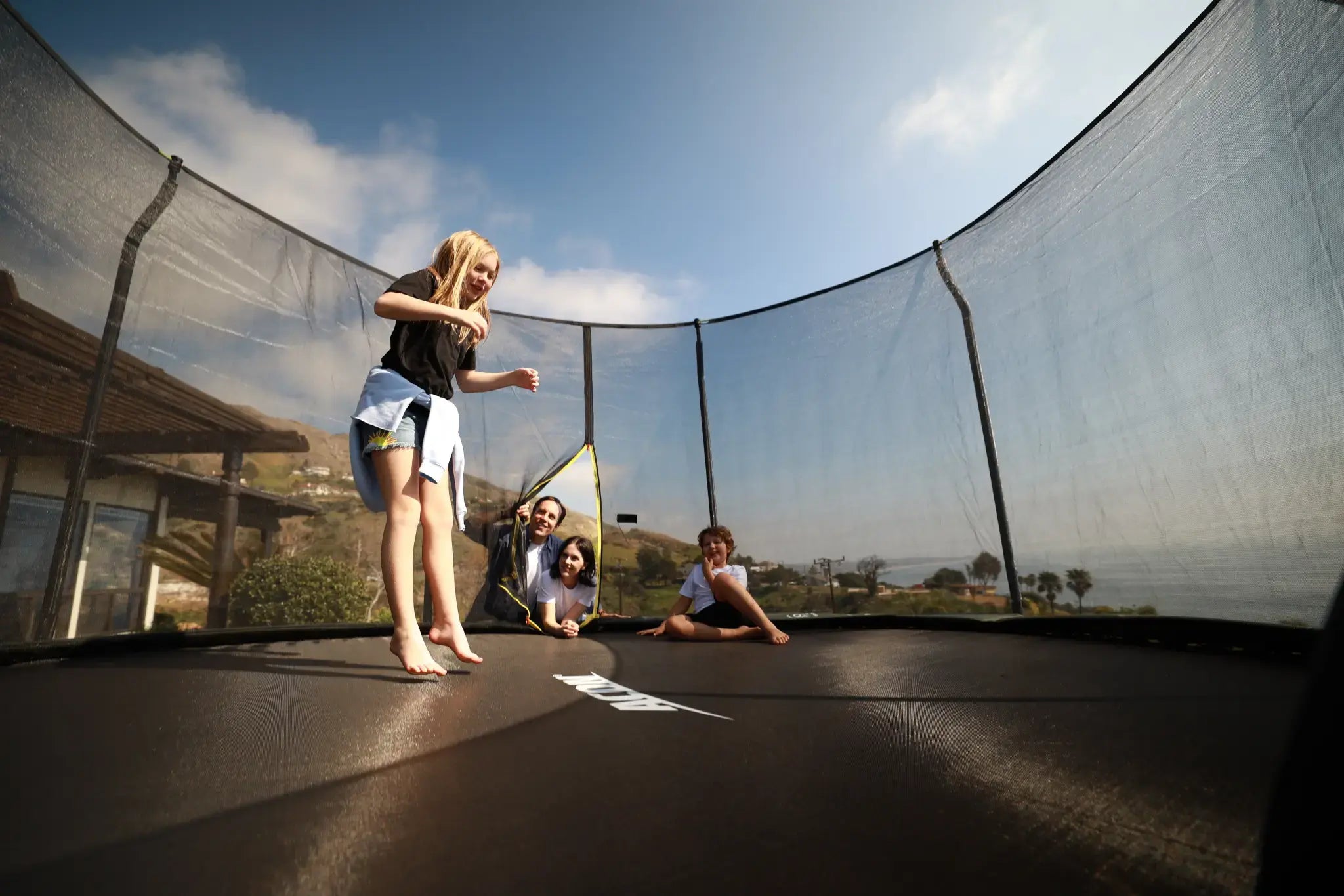 Girl jumping on an Acon trampoline surrounded with a safety net, with more people watching outside the trampoline.