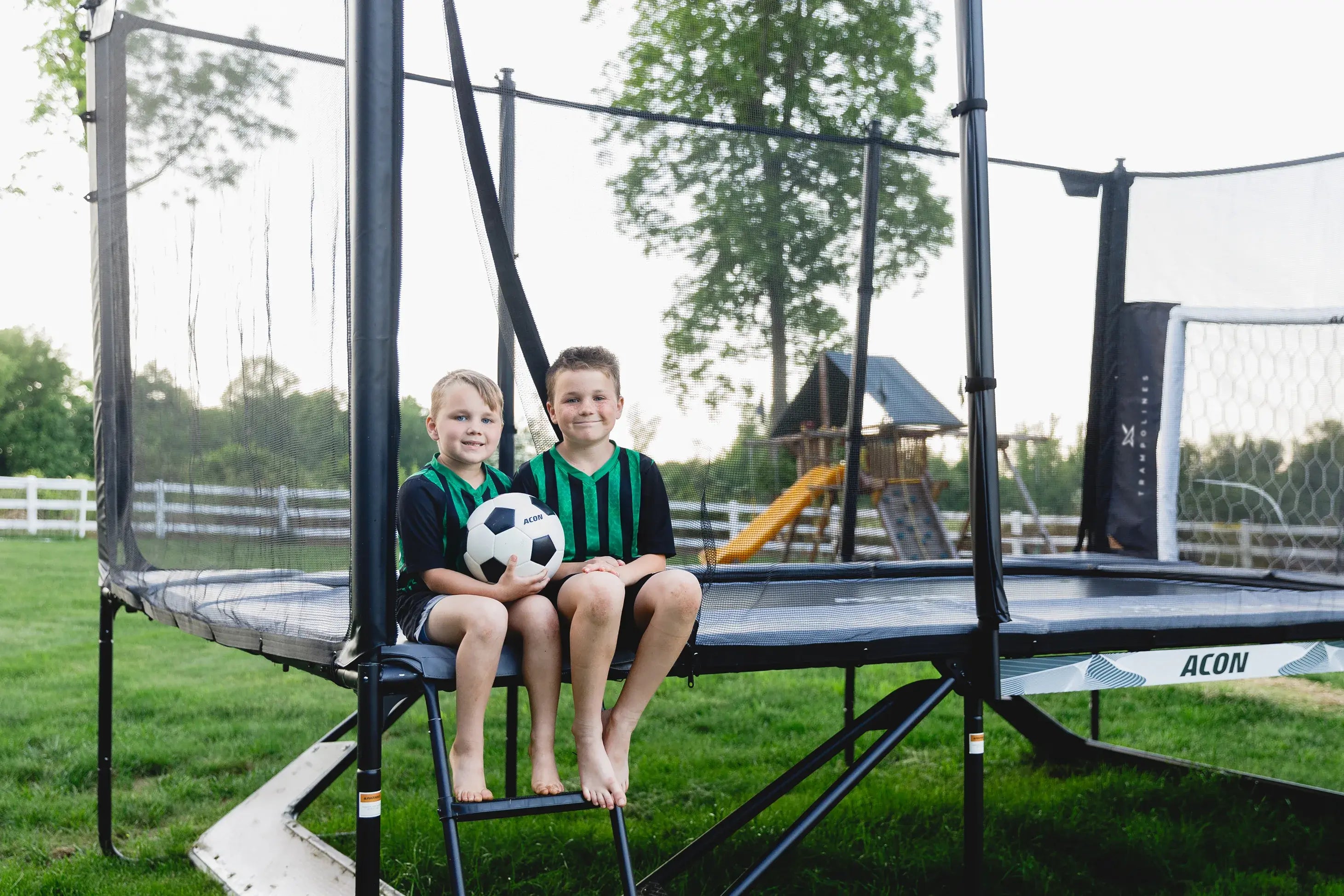Two kids with soccer ball on Acon trampoline in backyard with safety net and Acon Trampoline Soccer Goal.
