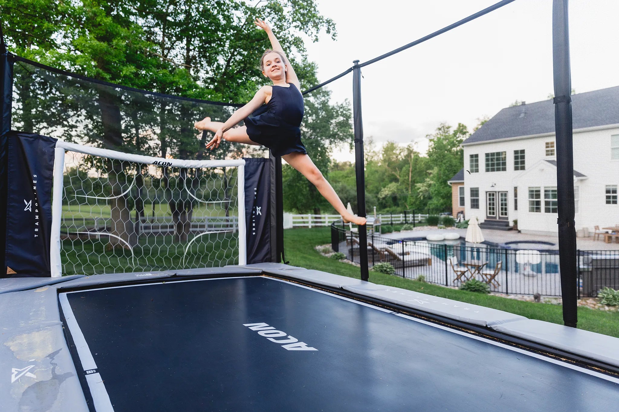 Girl doing a gymnastic jump on a Acon X trampoline with safety enclosure and soccer goal. 