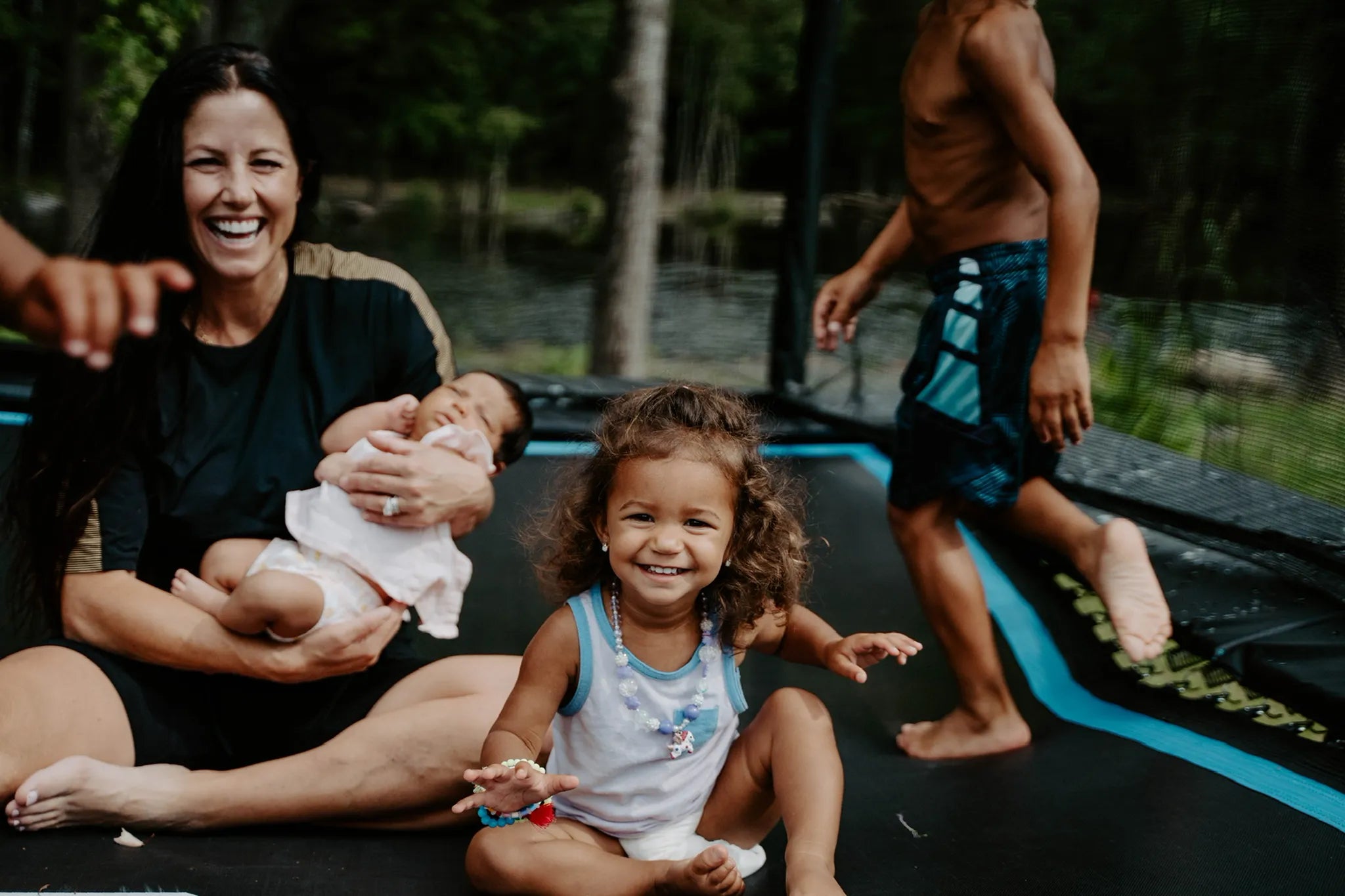 Mom holding a baby, one child sitting and third one running inside of Acon rectangular trampoline with safety net. 