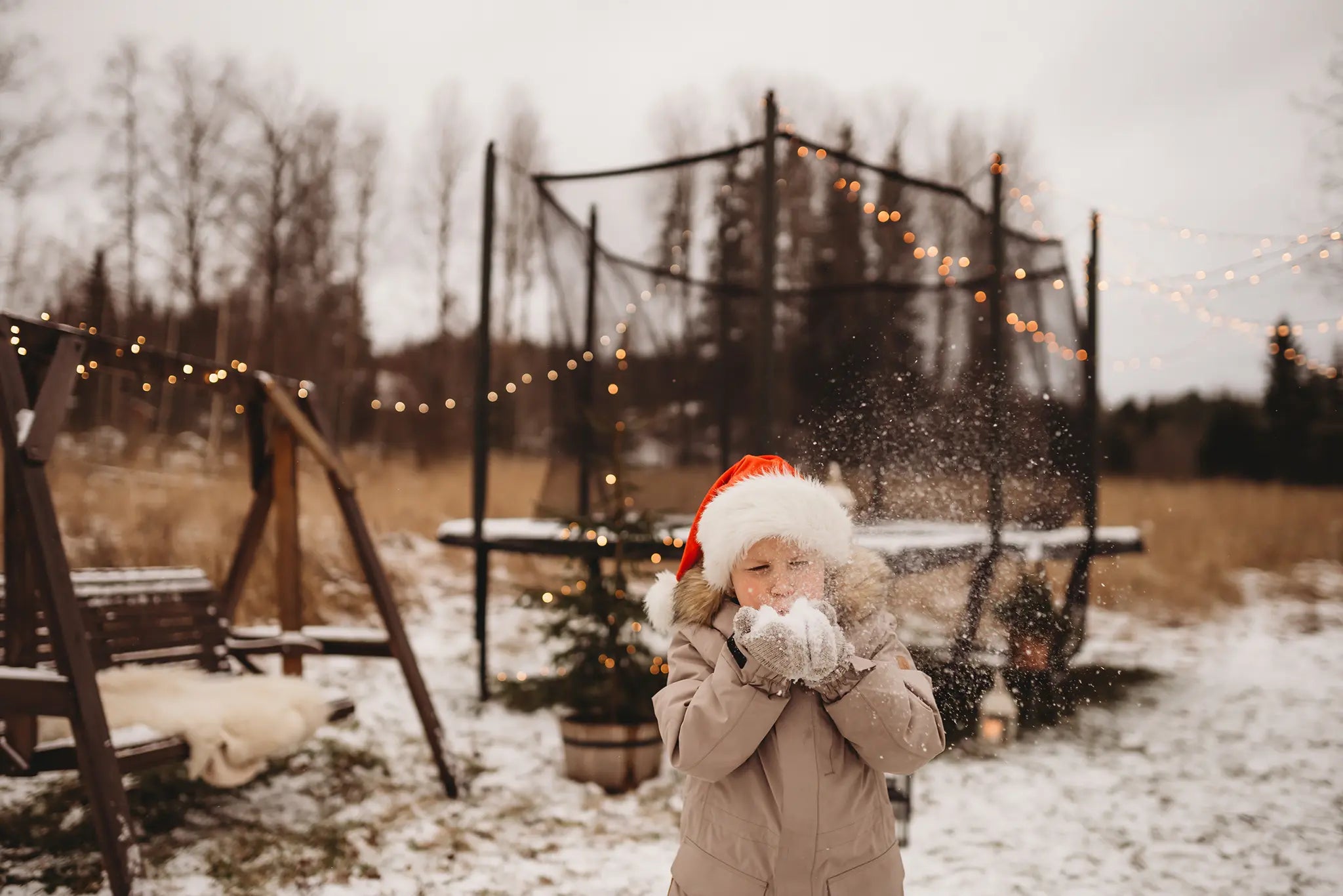 Young boy with santa hat blows snow in front of Acon trampoline, that is decorated with lights. 